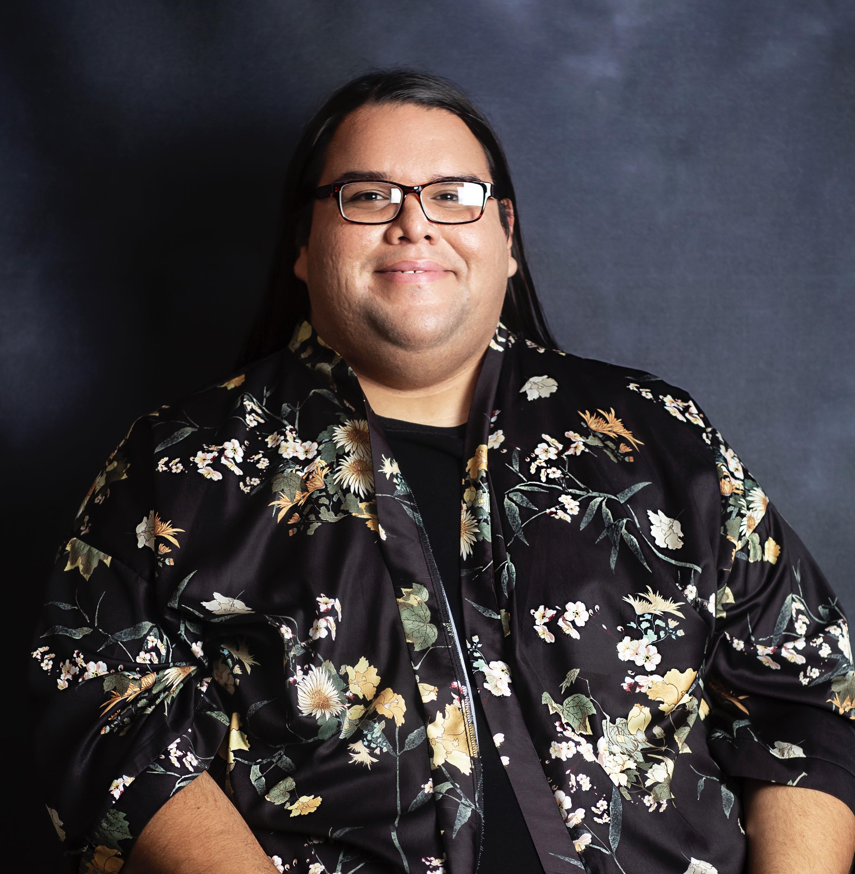 Headshot of a man smiling at the camera, with long hair, black frames, and wearing a shirt with flowers with a t-shirt underneath.