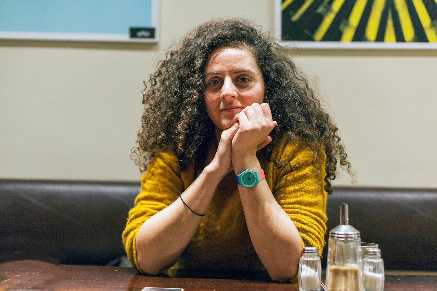 Portrait of woman with long curly hair wearing a mustard yellow sweater and aqua blue and pink watch. She is sitting in front of a table with her elbows on it. On the side there are salt, pepper and sugar shakers and behind some posters all which are out of focus.