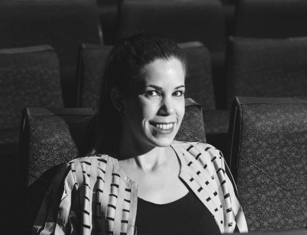 A black and white portrait of a woman sitting in a movie theater and smiling at the camera.