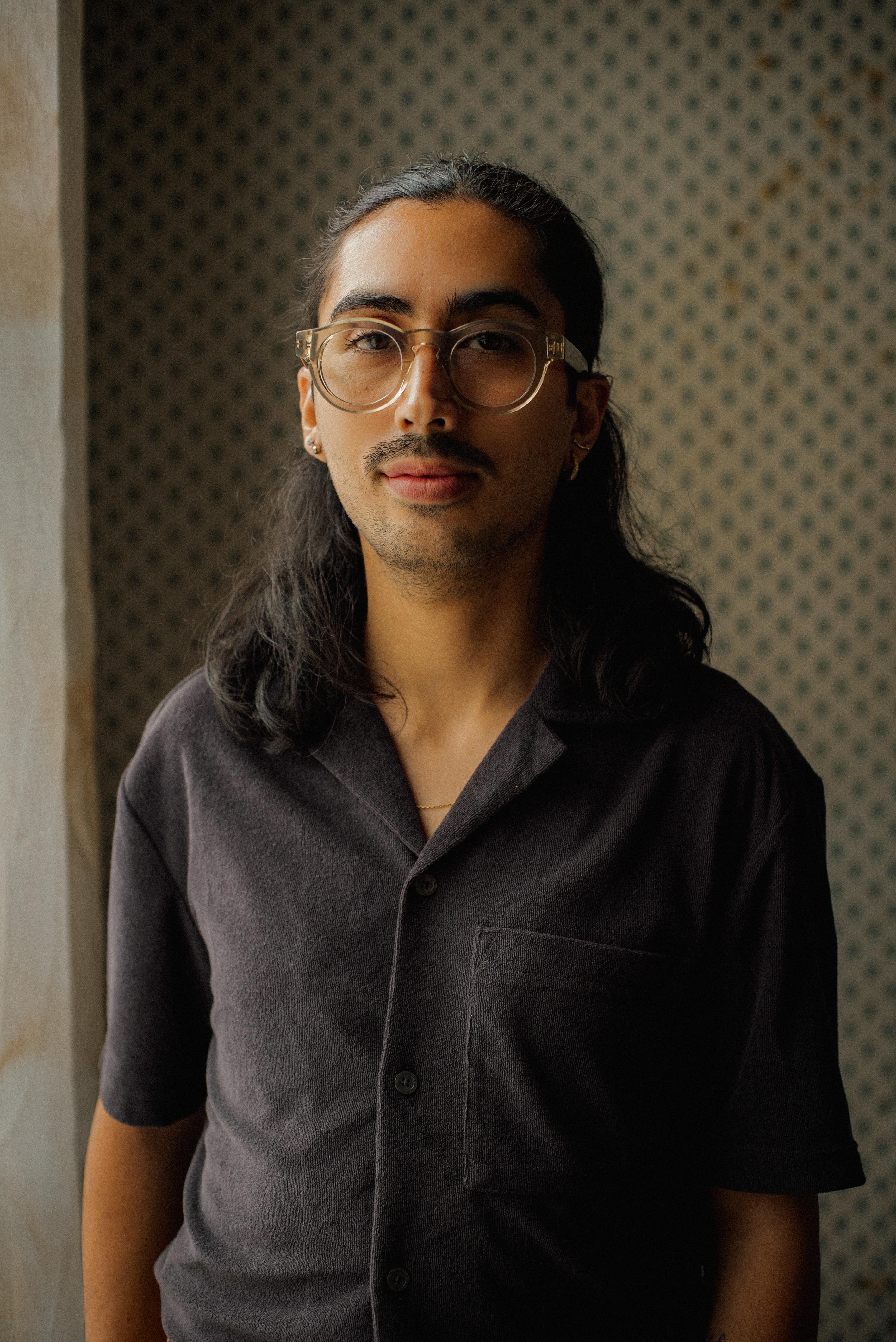 Headshot of Picho Garcia, a man with medium length wavy hair, mustache, and thick rounded frames.