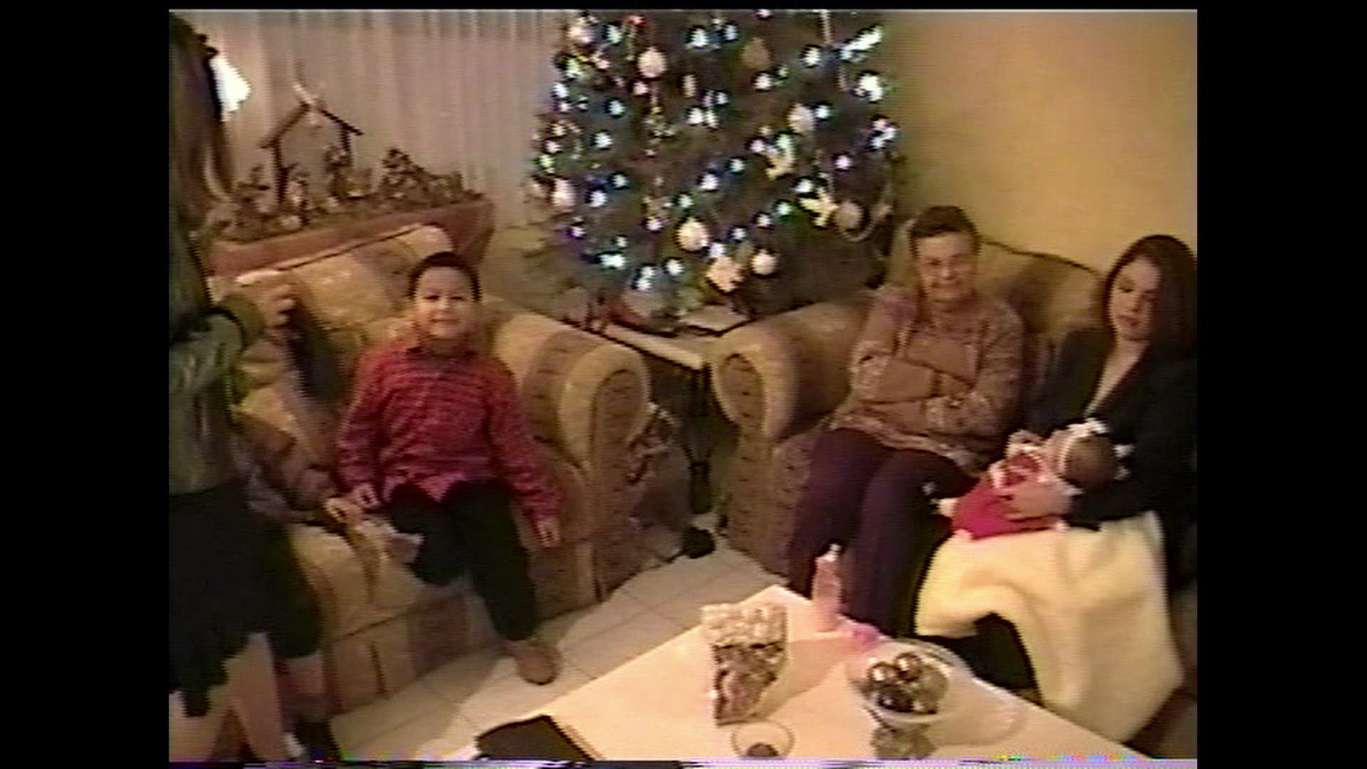 Still from #Mickey. Two women, one holding an infant, sit on a couch on the right. A child sits on a couch on the left. There is a Christmas tree and a Nativity scene in the background.