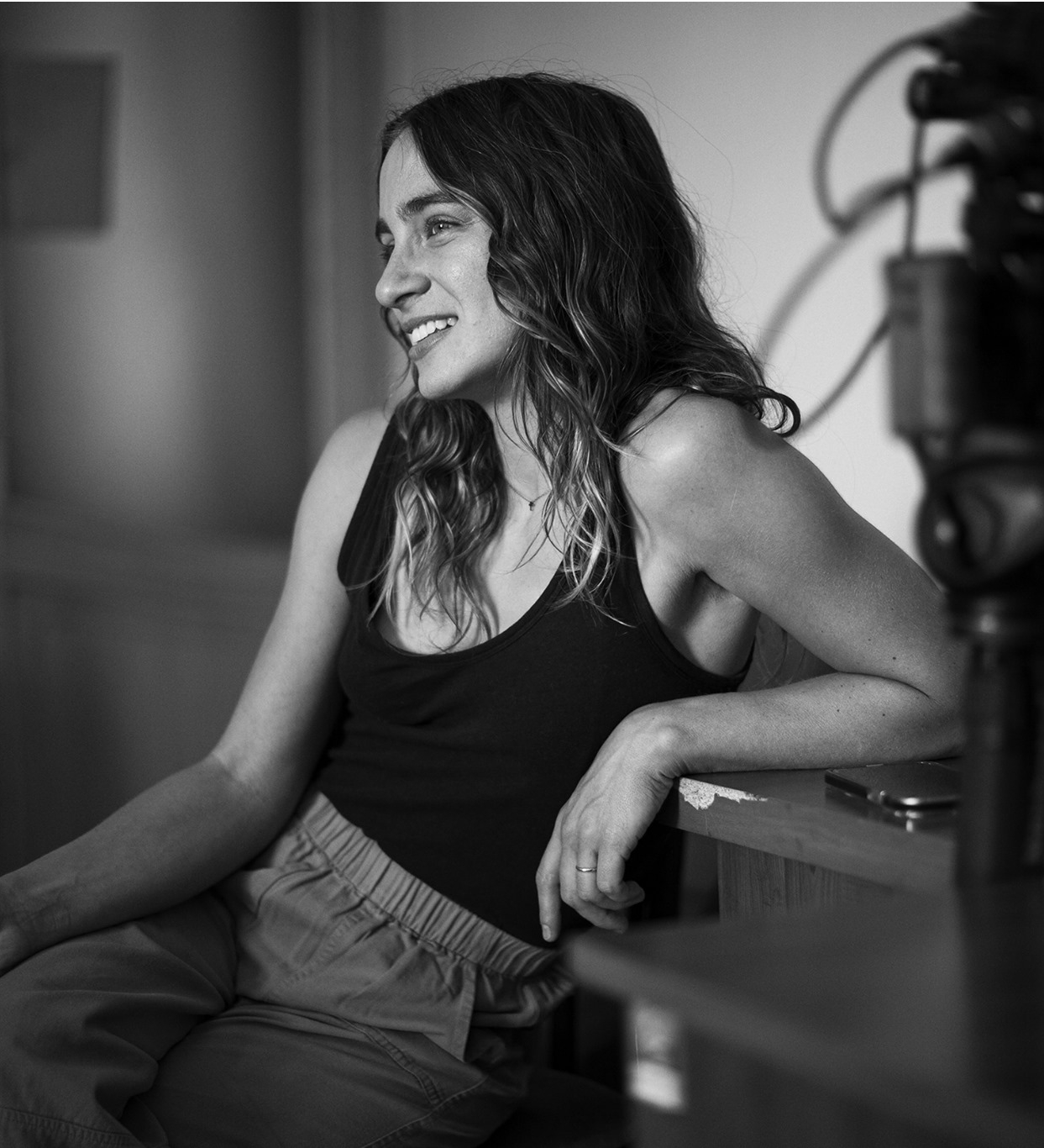 A black and white photo of a seated woman with curly dark hear, wearing a black tank top, seen from the side with a camera in the foreground.