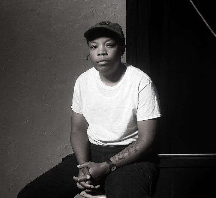 Portrait of a person sitting wearing a hat, white shirt and black pants.