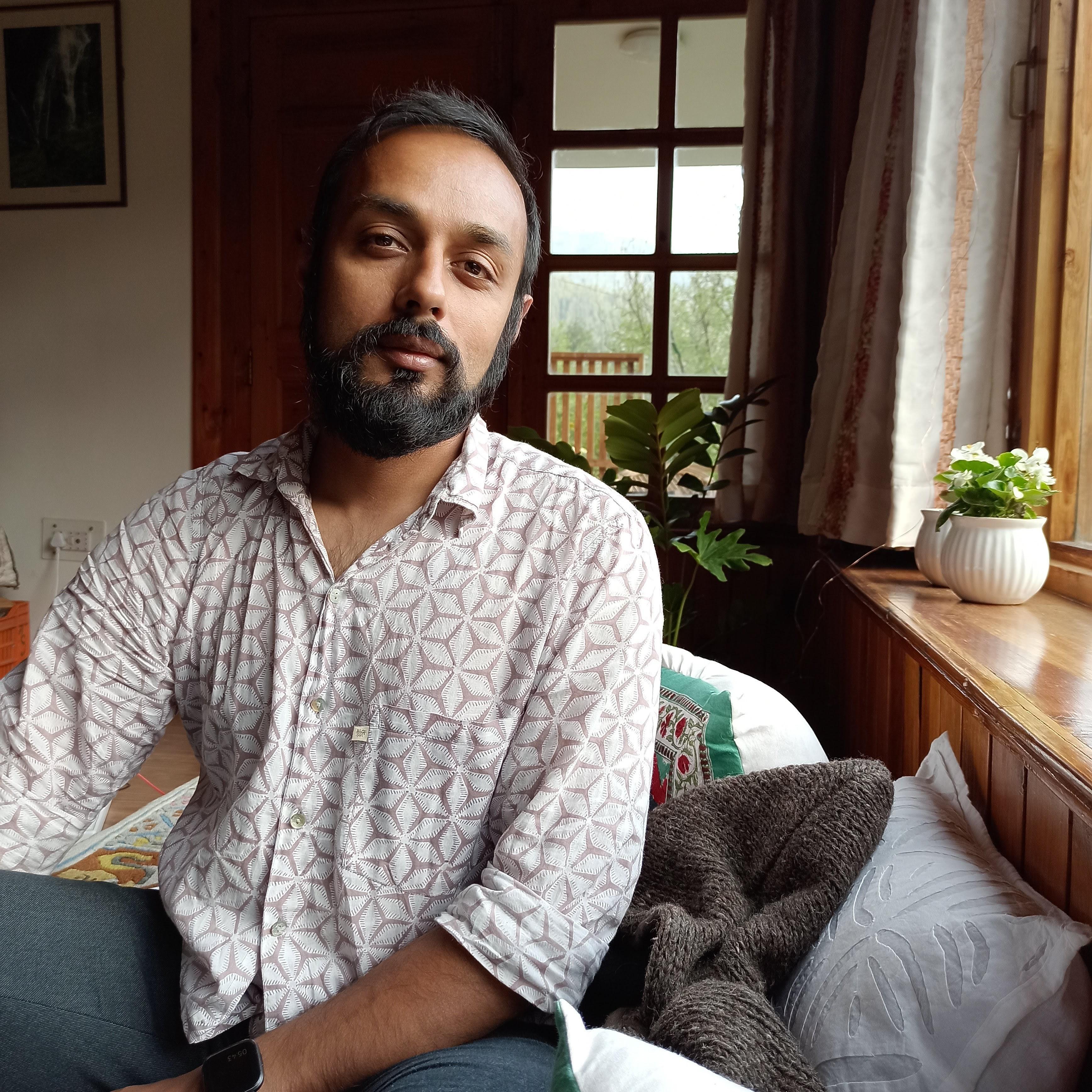Headshot of a South Asian man with a beard, wearing a light coloured shirt sitting in a room with wooden panels.
