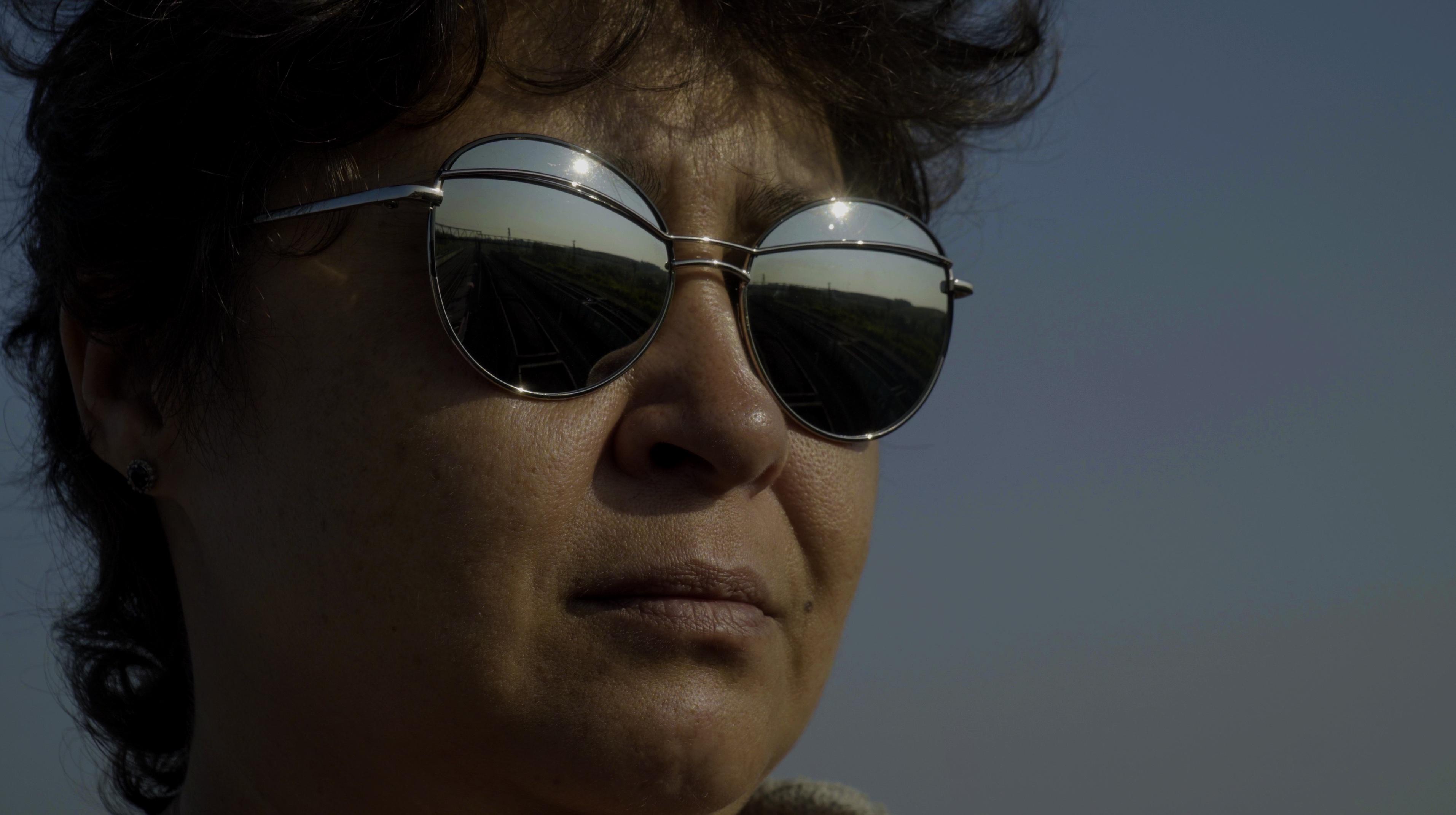 Still from Black Snow. A head-only view of a woman with short, brown hair wearing mirrored sunglasses with coal trains reflected in them.