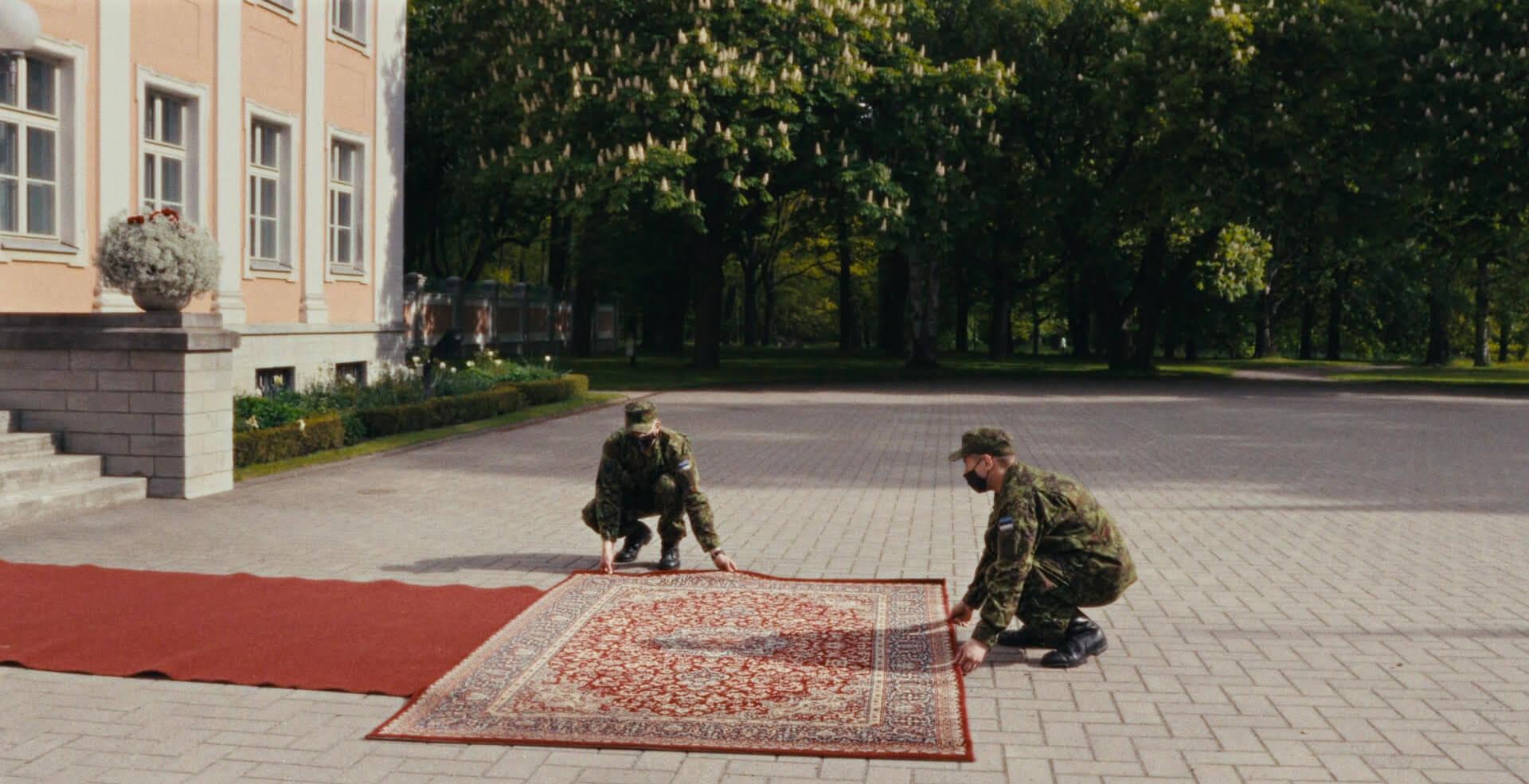 Still from Not Made For Politics. Men in military uniforms straighten a rug in a driveway.