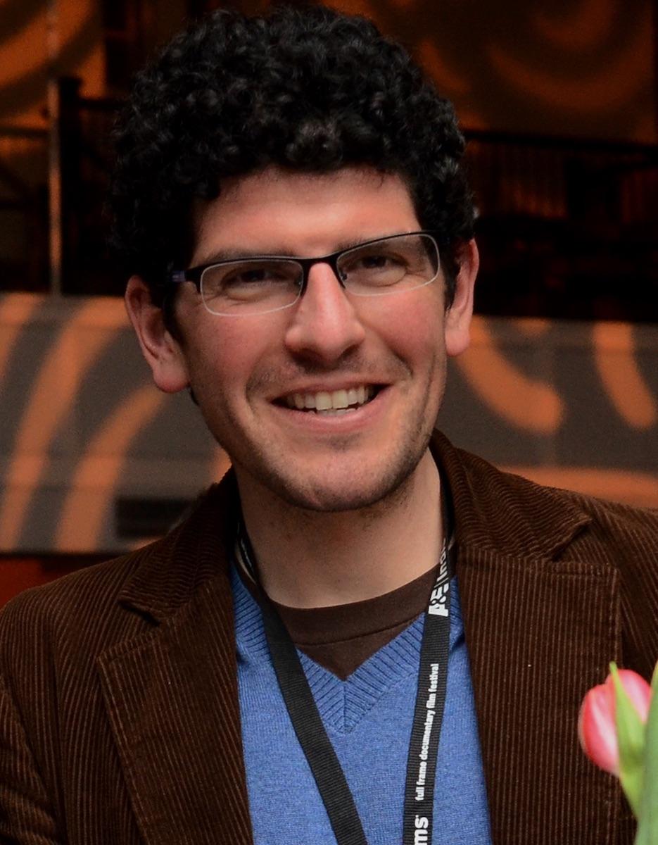 Mike Attie looking directly at camera. He is wearing eyeglasses and has a festival lanyard around his neck.