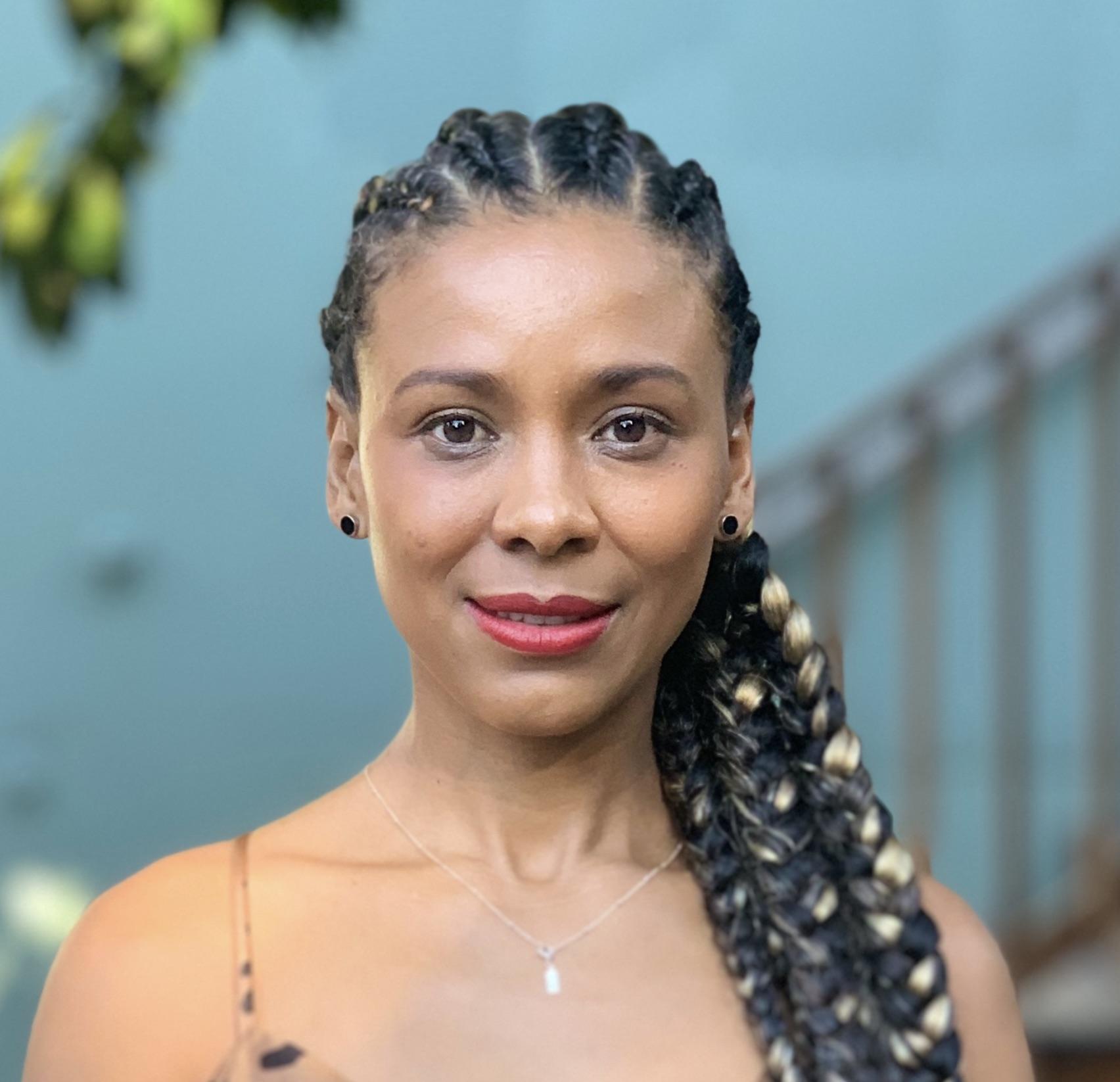Débora Souza Silva looking directly at the camera. She has braided black hair and golden highlights.