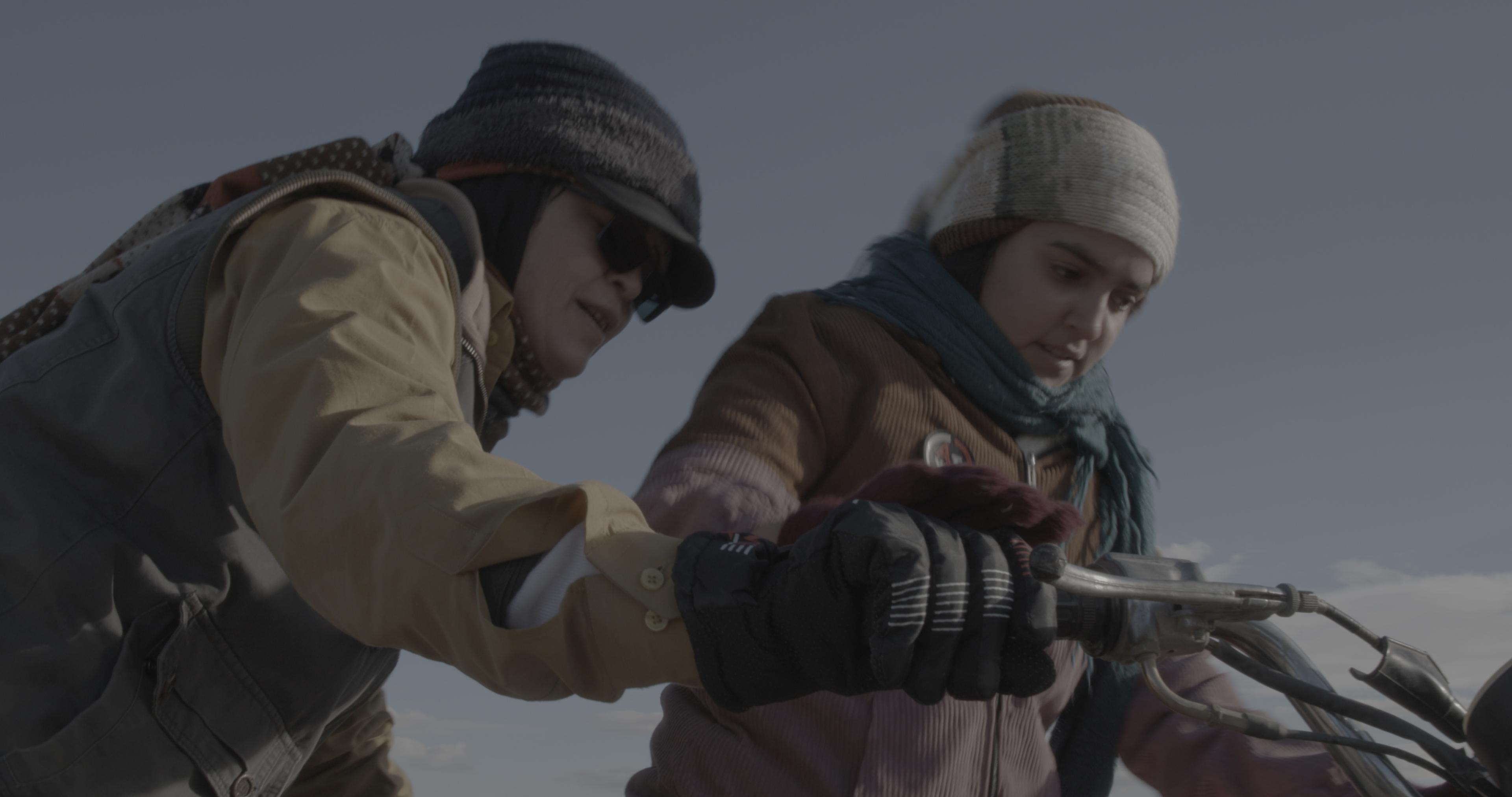A low angle photo of a young girl starting a motorcycle. A woman stands behind and guides her.