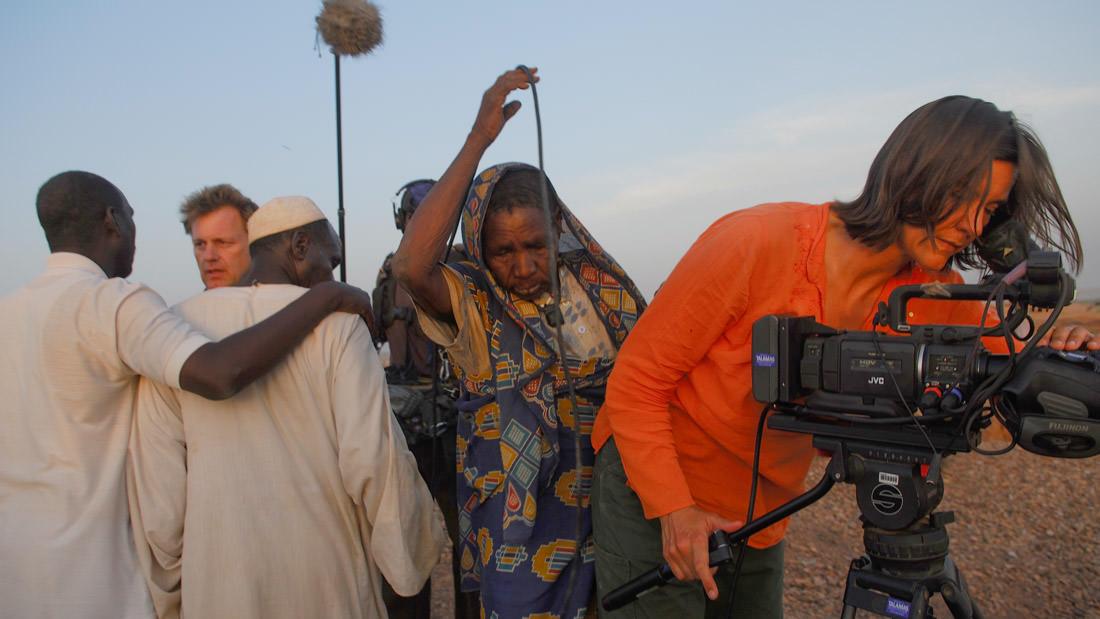 A woman wearing a bright orange shirt sets up a camera while a group of documentary subjects gather around a two crew members.