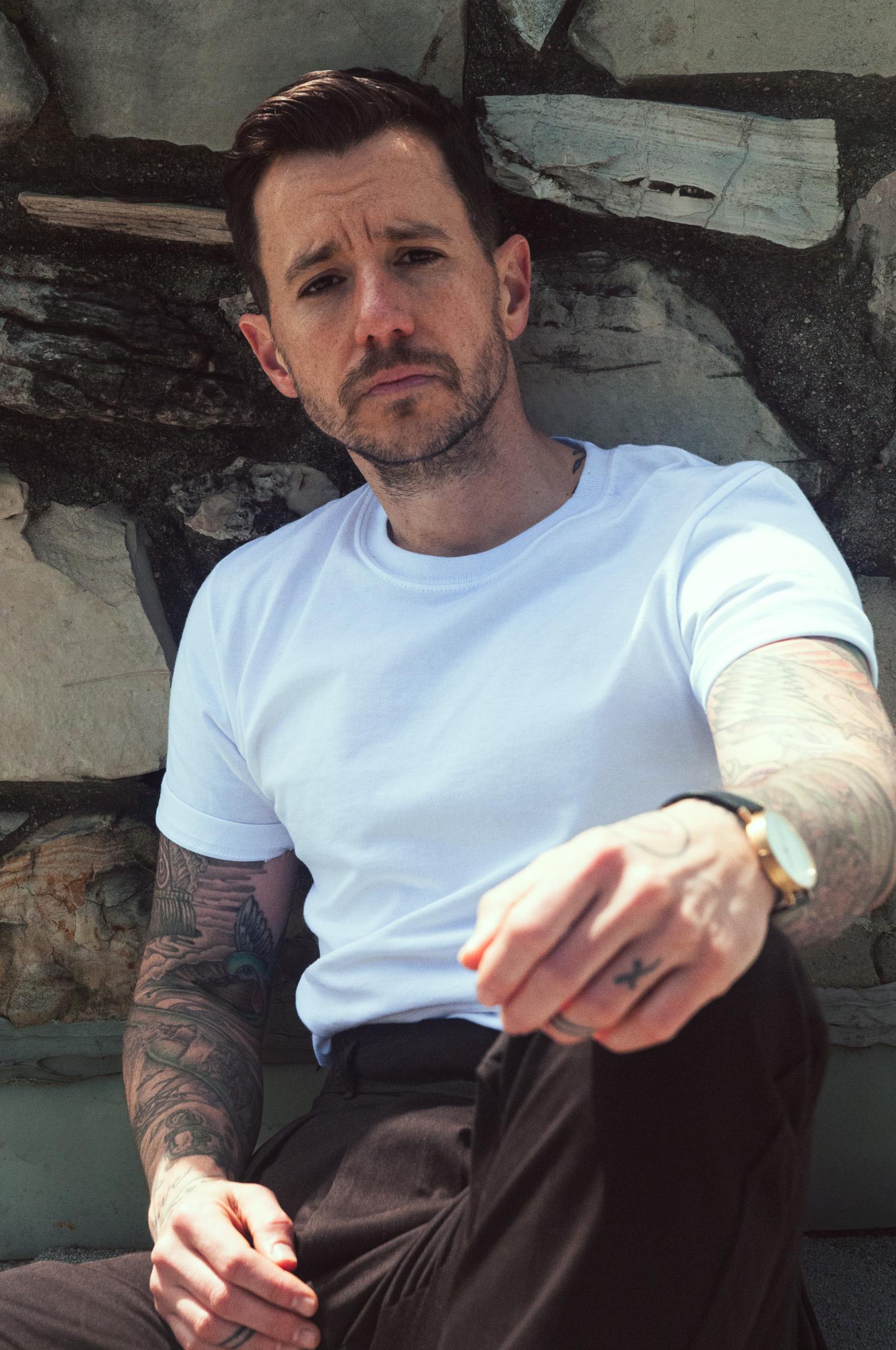 A man sits against a stone backdrop and looks into the camera with a serious expression.