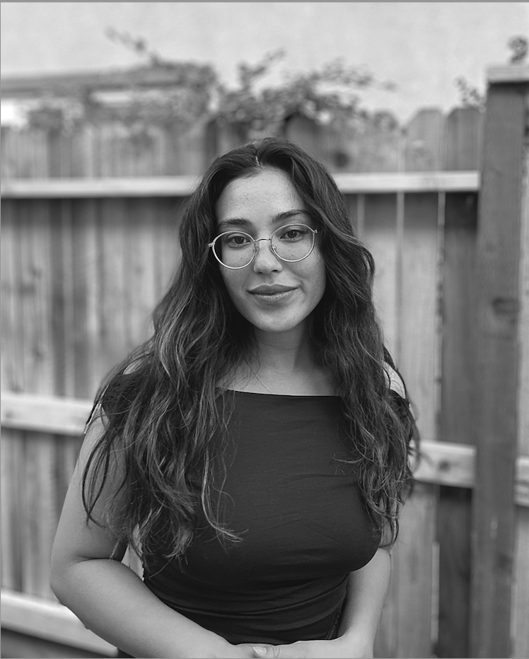 A mixed race woman stands in front of a wooden fence. She has long, dark hair and wears wire rimmed glasses and smiles slightly while looking directly at the camera. A headshot for filmmaker Bree Nieves Robert.