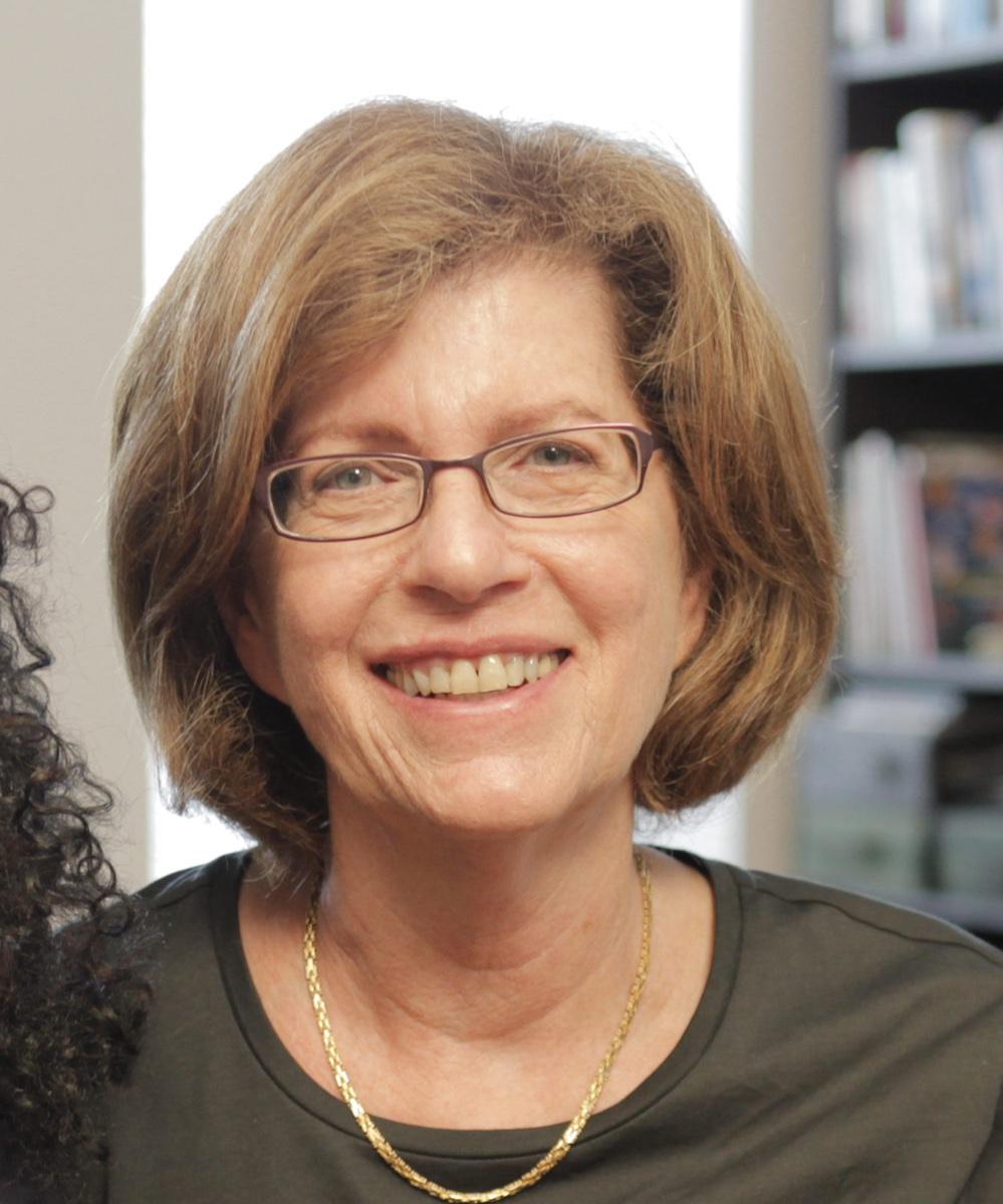 Barbara Attie looking directly at camera. She is wearing eyeglasses and a gold necklace. The background of bookcases is out of focus. Black and white portrait.