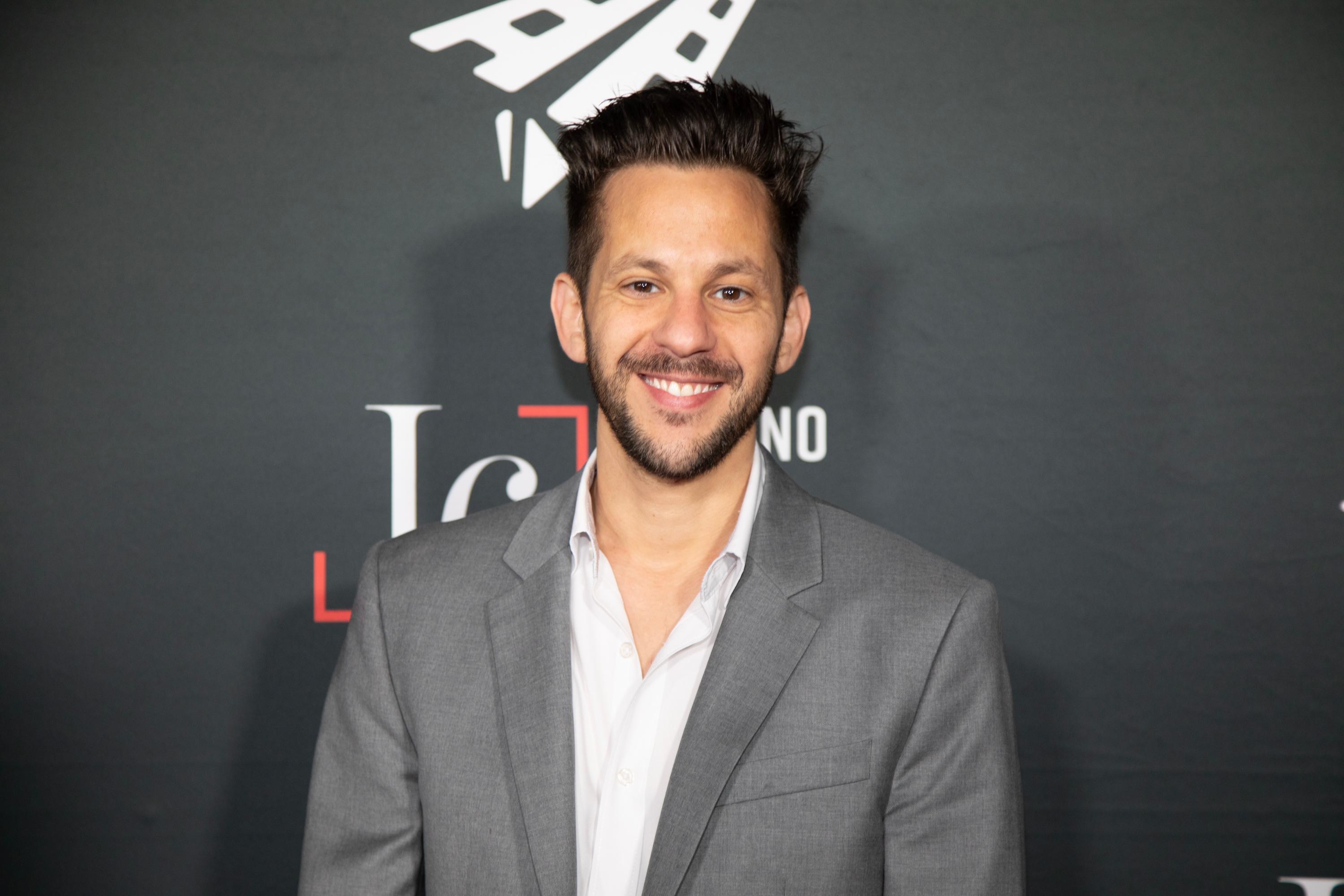 A man in a suit stands in front of a step and repeat banner and smiles.