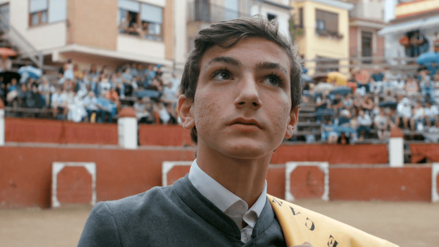 A still from The Boy and the Suit of Lights. A close-up shot of a young boy wearing a grey suit and a yellow cloth draped over his left shoulder. Behind him, a crowd of people sits in stadium seating. He has short brown hair and brown eyes.