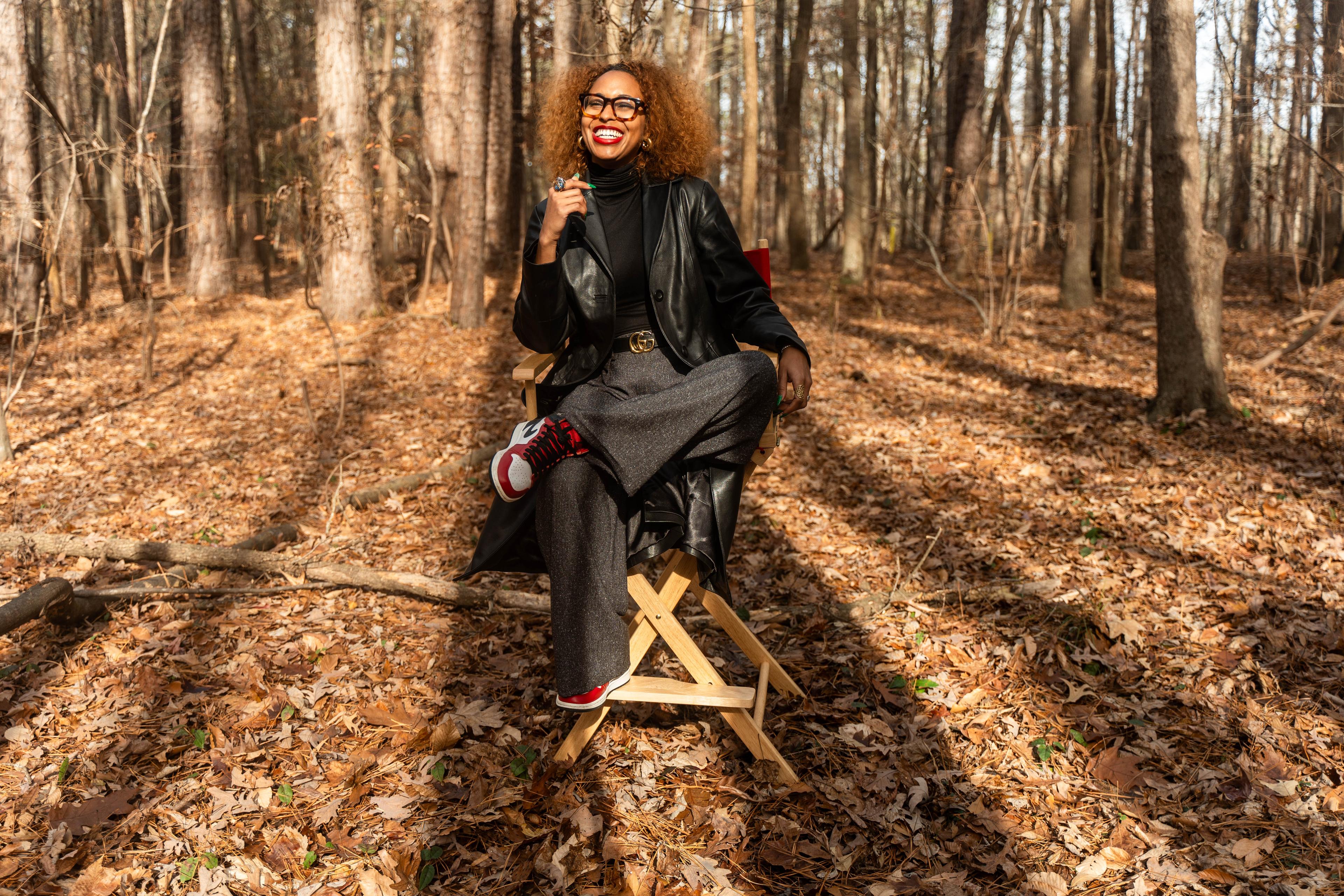 Brown skinned woman with red lipstick and brown glasses smiles in the woods surrounded by trees.