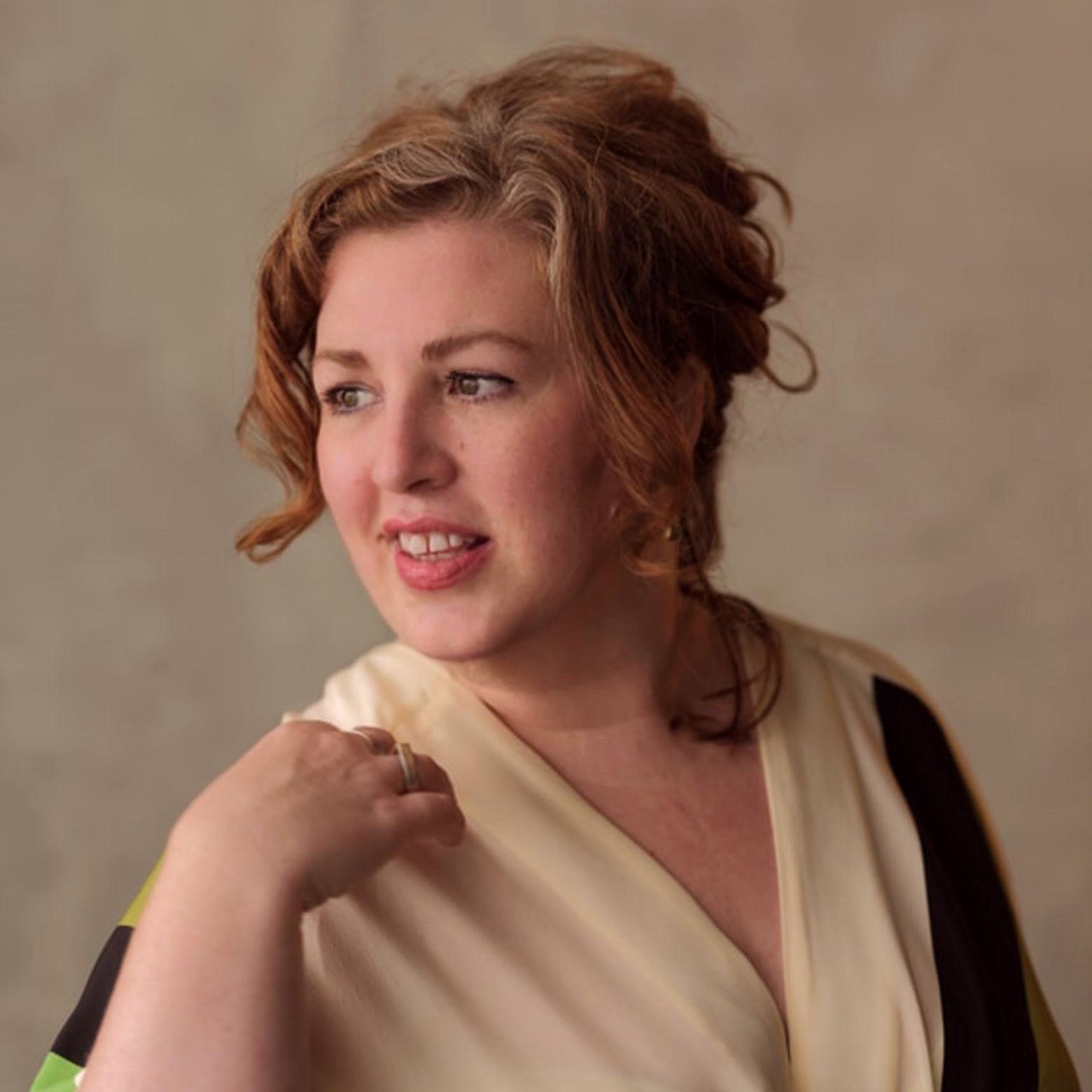 Vertical portrait of a woman looking off frame smiling. She is wearing v neck beige, black and green blouse. Her hair is done up and she has ginger hair.