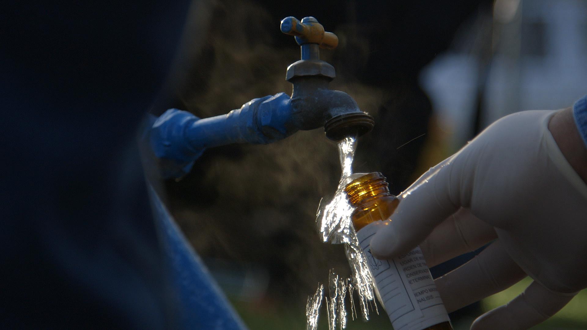 Still from The Age of Water. Somebody wearing gloves pours water from a tap into a small bottle.