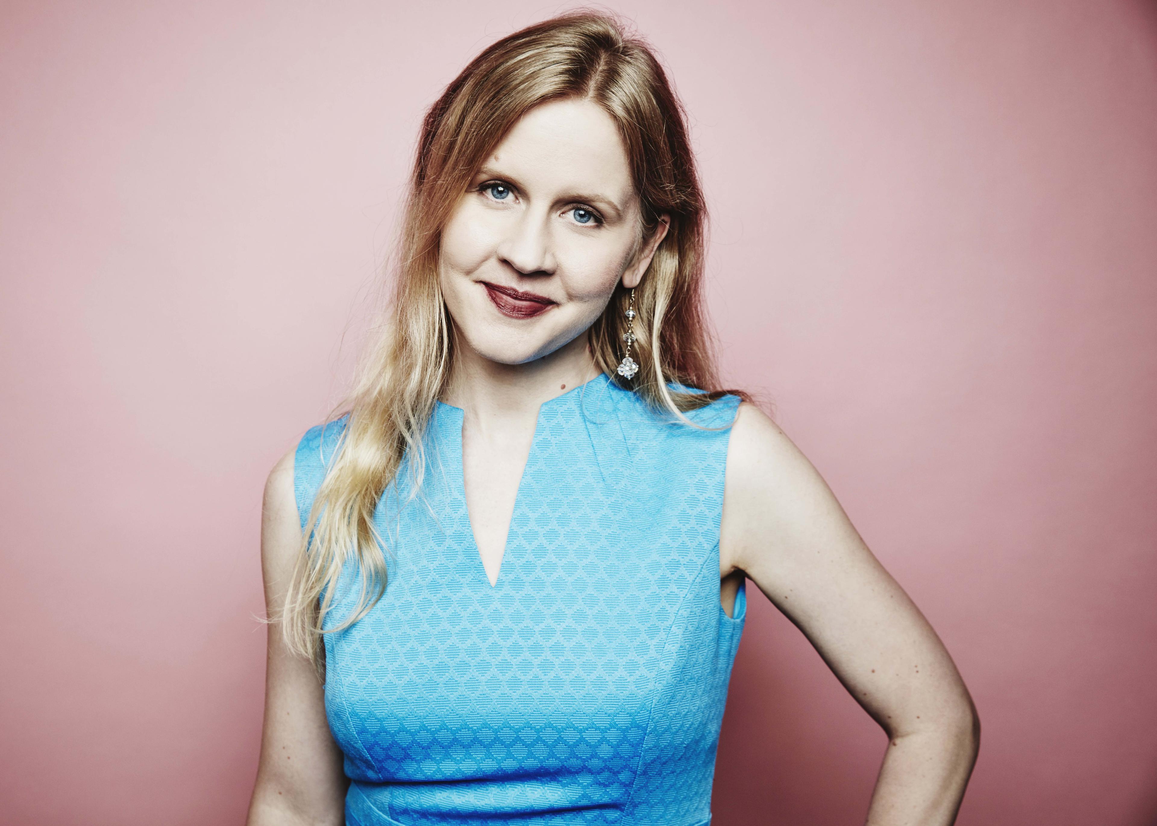 Justine Nagan Smiles at the camera with her head slightly tilted. She is wearing a blue dress and is standing in front of a pink background.