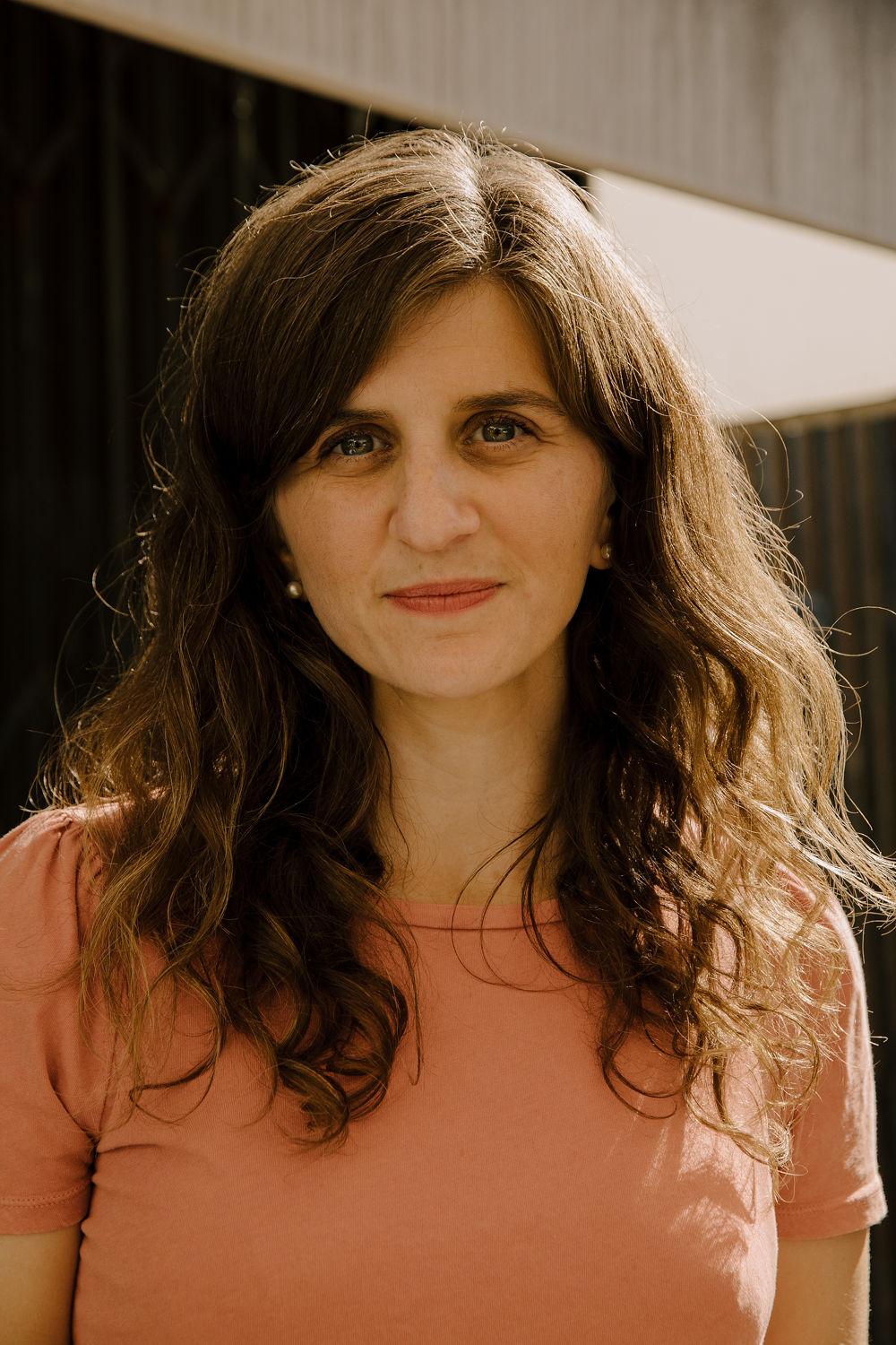 A white woman with long, brunette hair wearing an orange t-shirt.