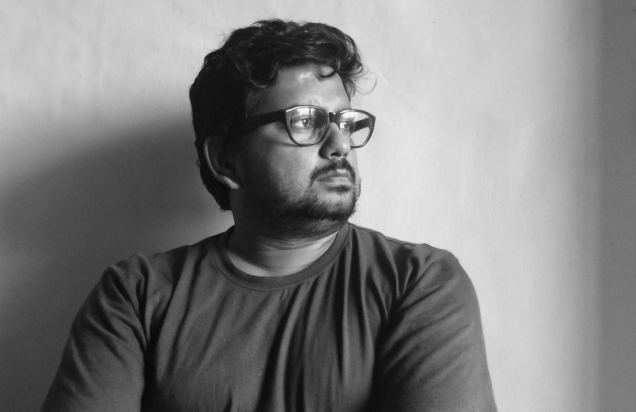 A black and white portrait of a man with dark hair and glasses, looking off into the distance.