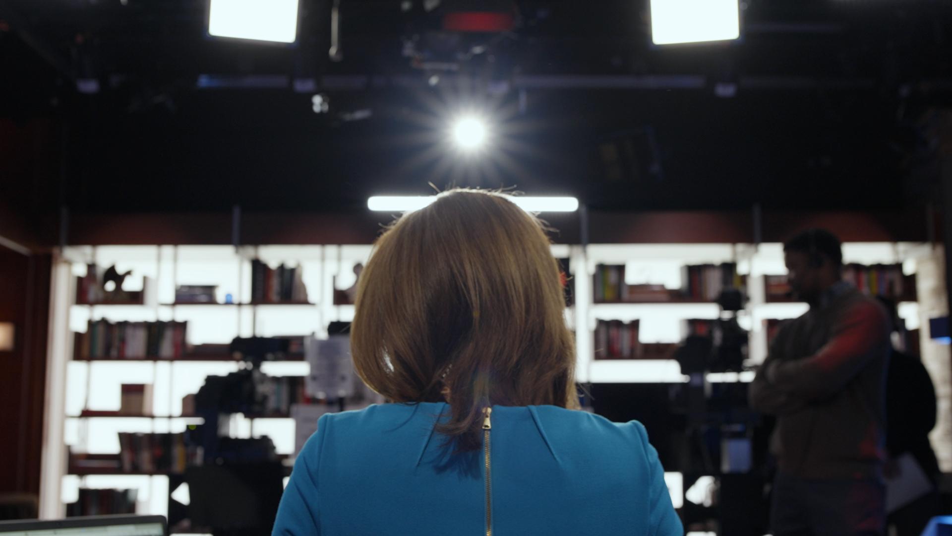 Angled from behind, a newswoman in a blue top is sitting, looking towards a camera and a bright light. Her POV emits a sense of pending action.