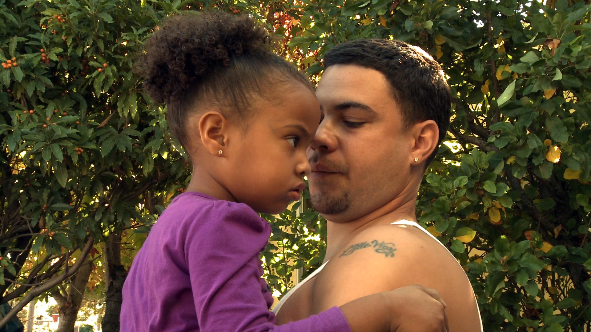 Still from The Surrender of Waymond Hall. Waymond Hall holding a young kid in front of shrubbery. Color photograph.