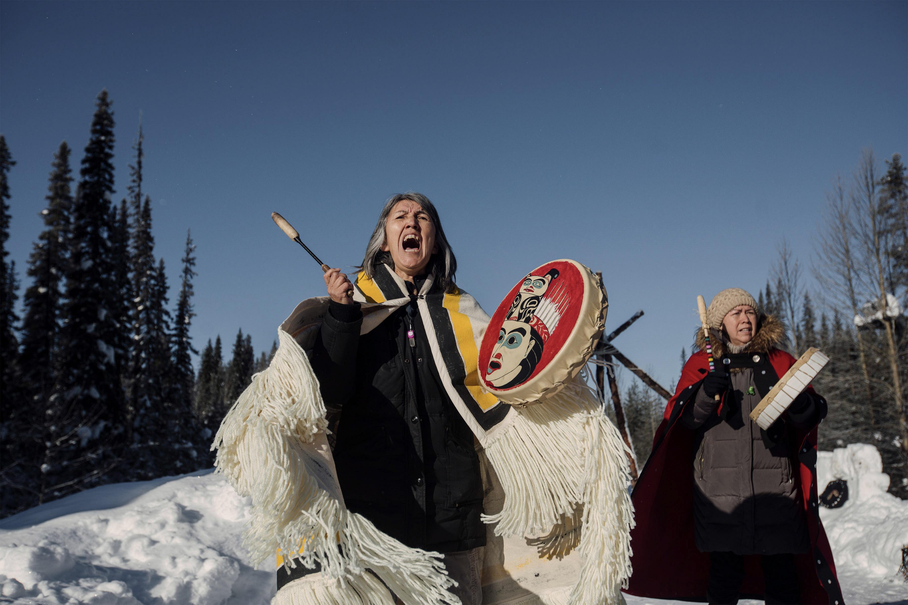 Still from Yintah. Two women sing and hit ceremonial drums against a snowy backdrop and blue skies.