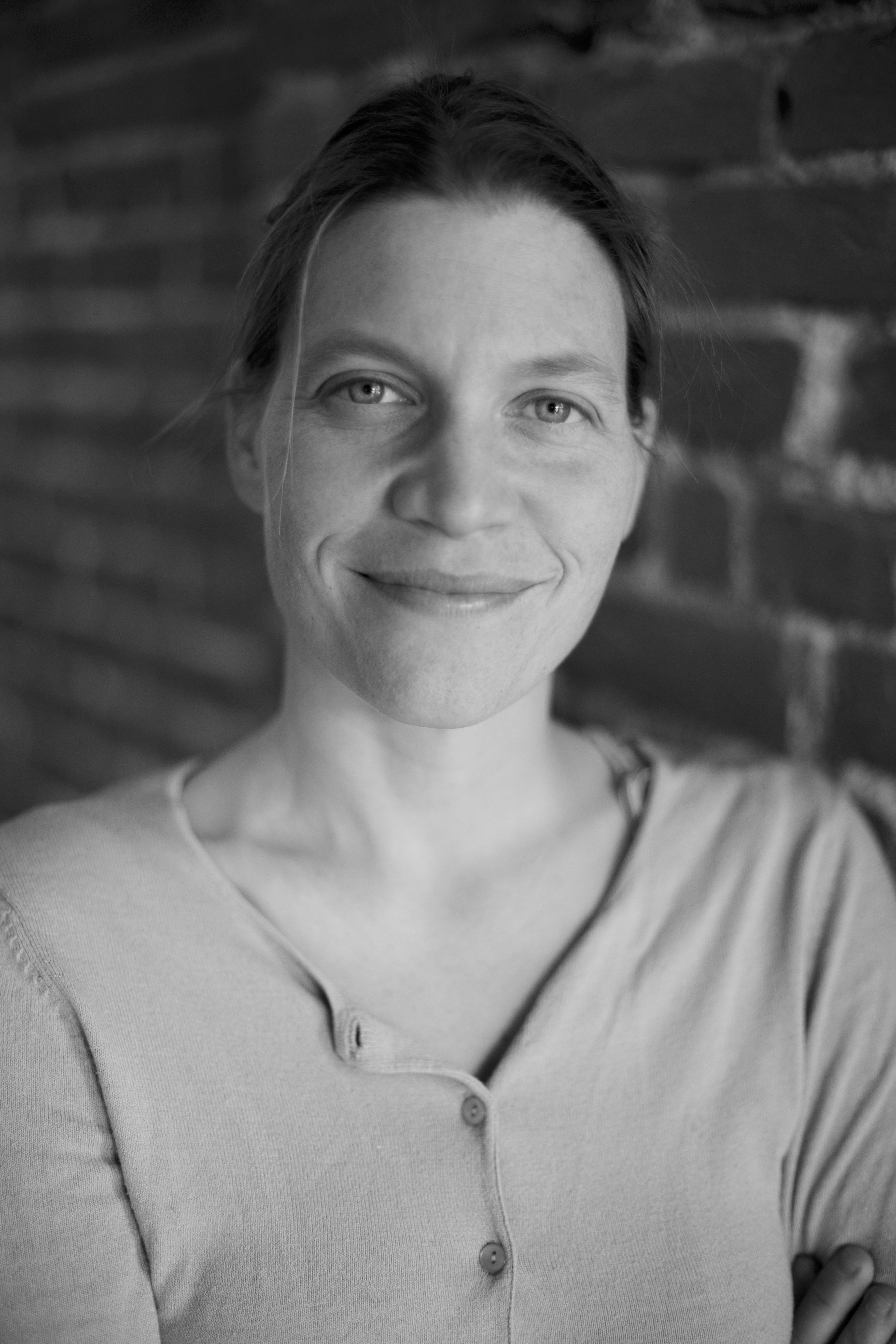 Emily Topper smiles at the camera with her hair pulled back and arms crossed. The photo is in Black and White.