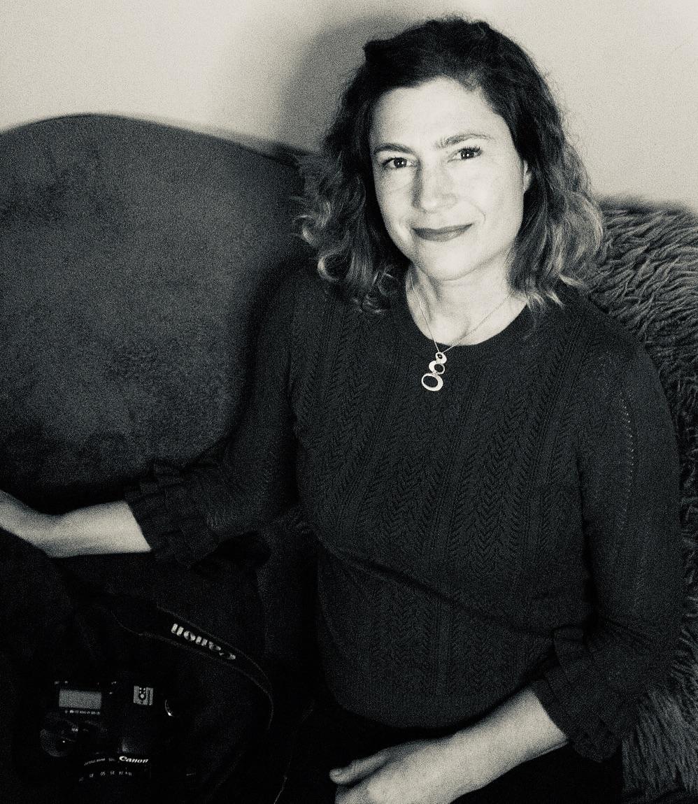 A black and white portrait of a woman sitting on a couch with a camera in her lap. She smiles into the camera.