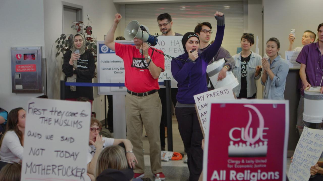 Still from An Act of Worship. People participating in a protest against the Muslim ban, the hold posters.