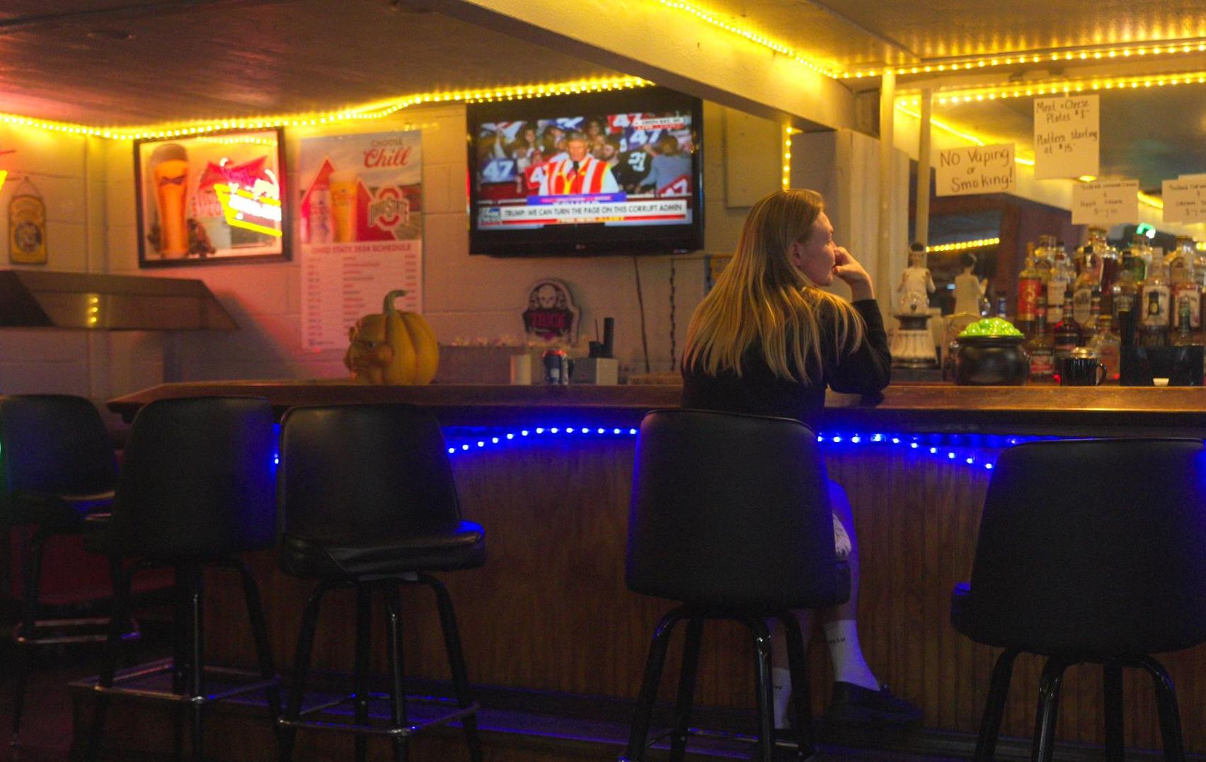 A woman sits alone at a bar while the news plays.