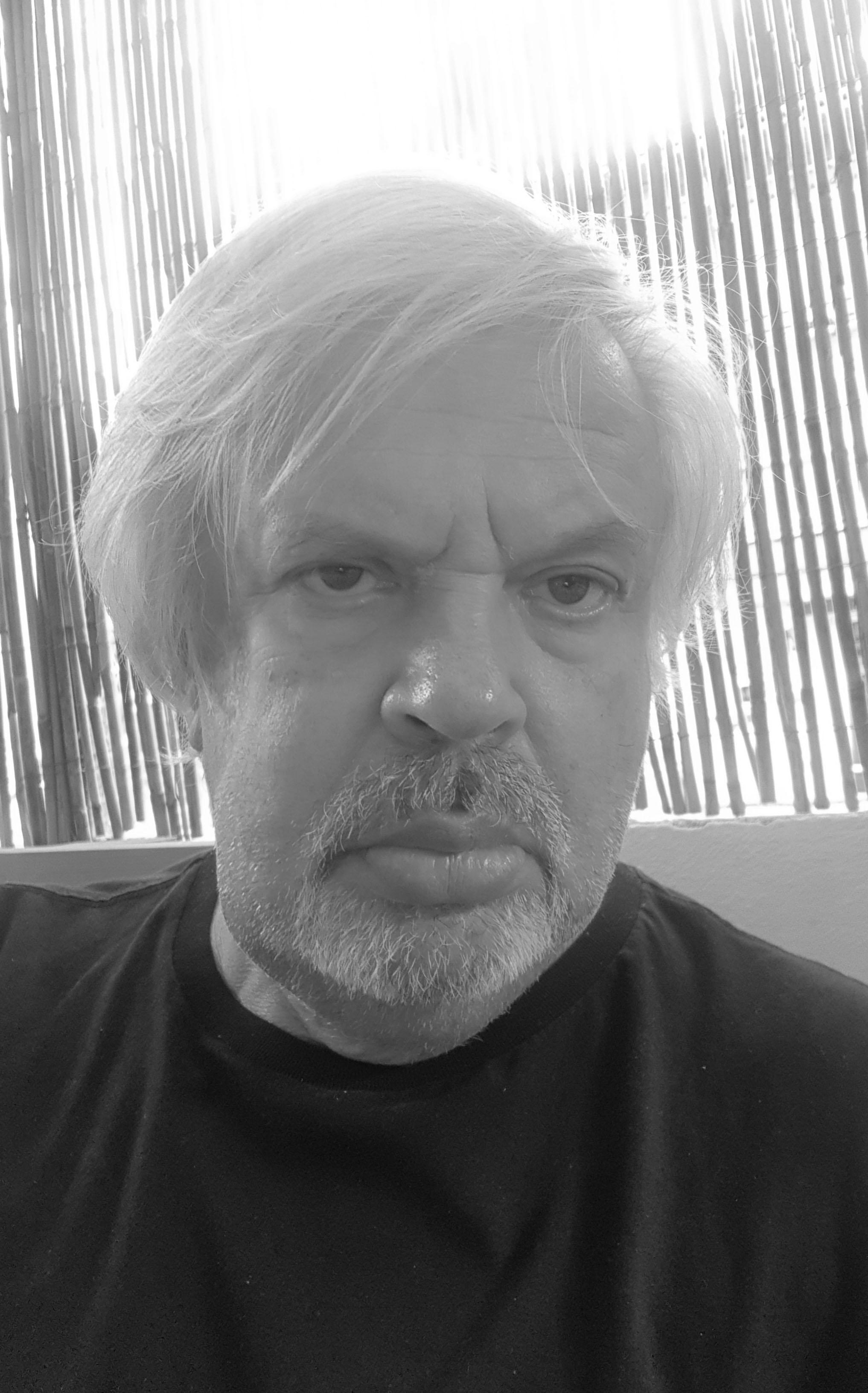 Valeryi Kalmykov looking directly at camera. He is wearing a black t-shirt and is sitting in front of bamboo shades. Black and white portrait.