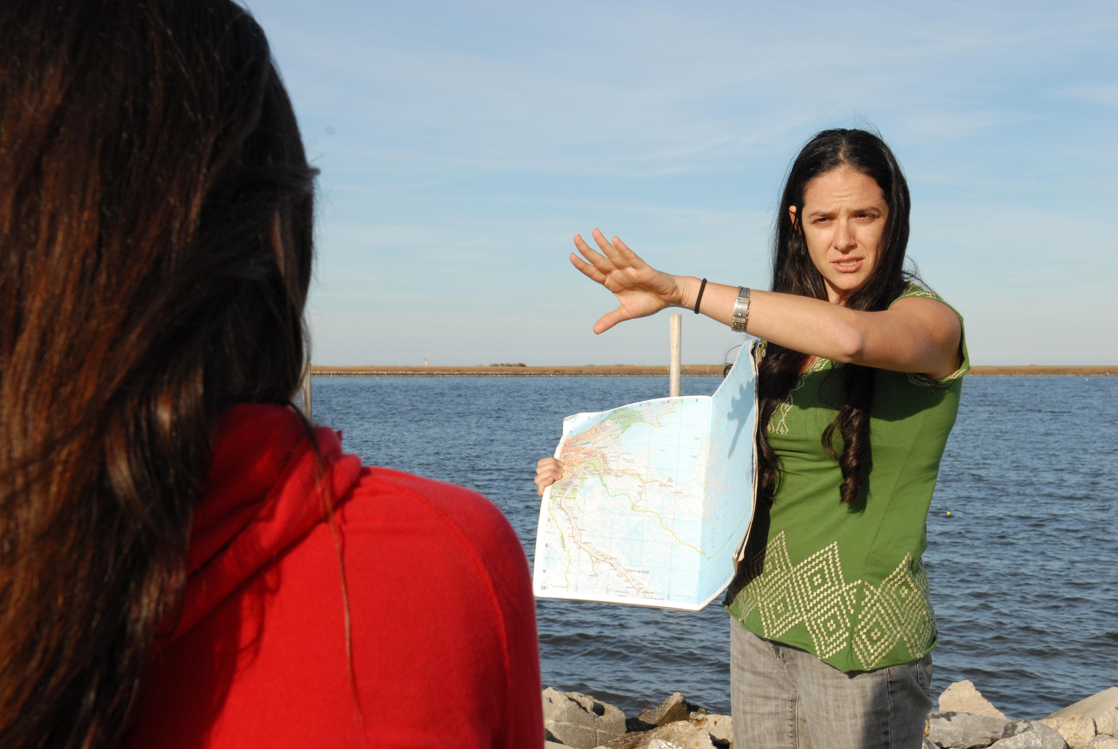 A woman stands in front of a beach and holds up a map. She holds her hand up as she speaks.