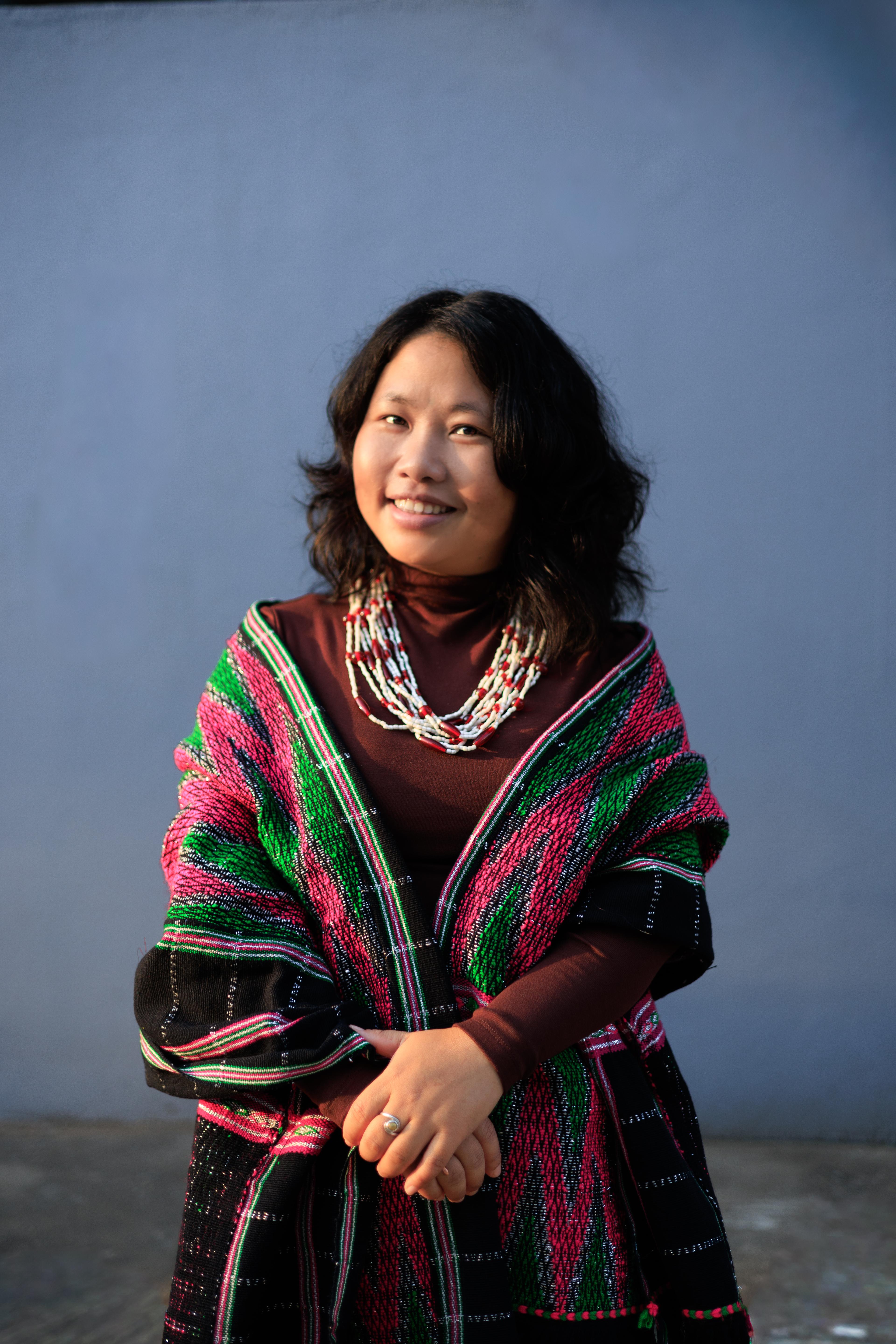 A young Miju Mishmi woman, with open hair, is standing in her traditional attire, smiling gently at the camera.