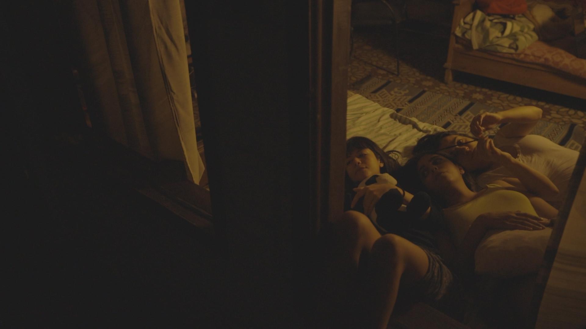 A still from House No. 7. Three young girls lay on a mattress in a dimly lit apartment.