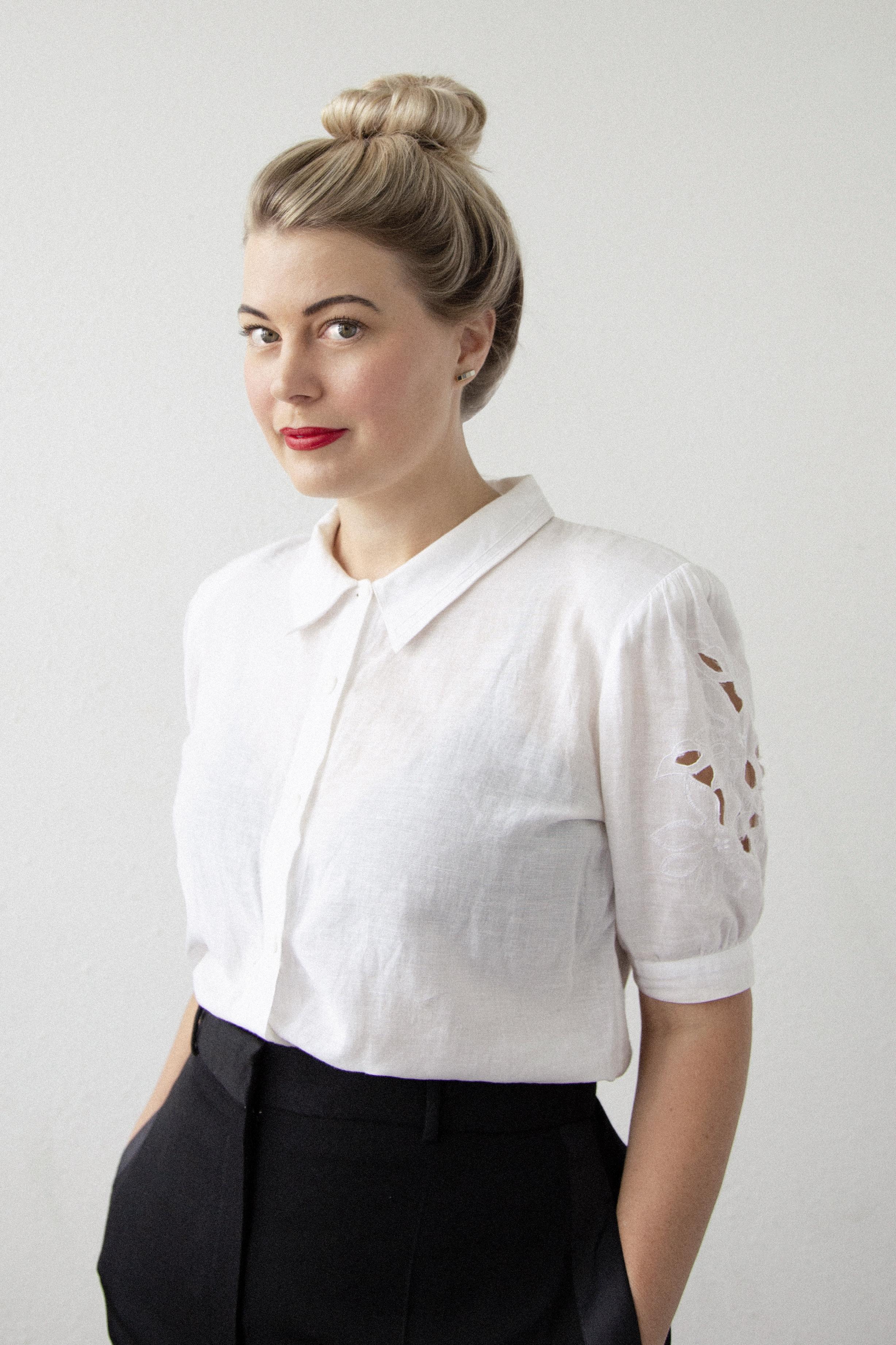 A woman stands in front of a white backdrop and looks into the camera. She has her hair in a bun and wears a white collared shirt.