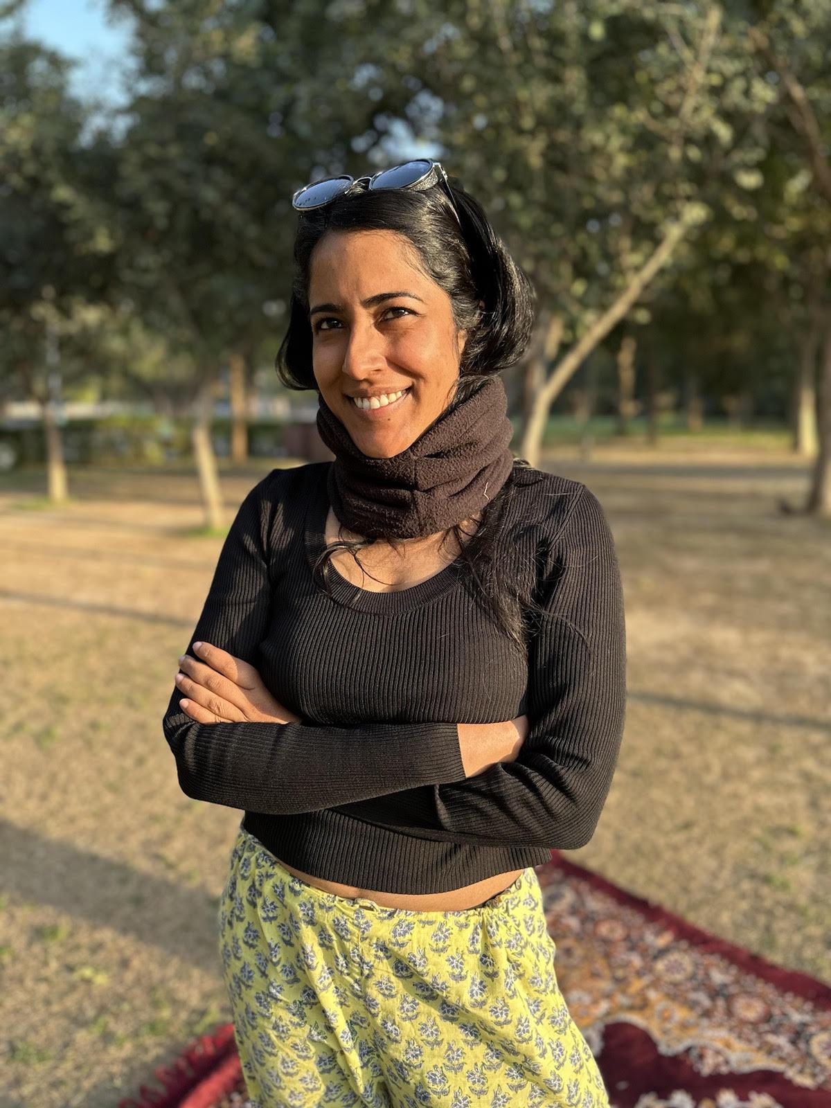 Headshot of a South Asian woman with black hair, wearing a black top and mustard-colored pants, standing against a background of trees.