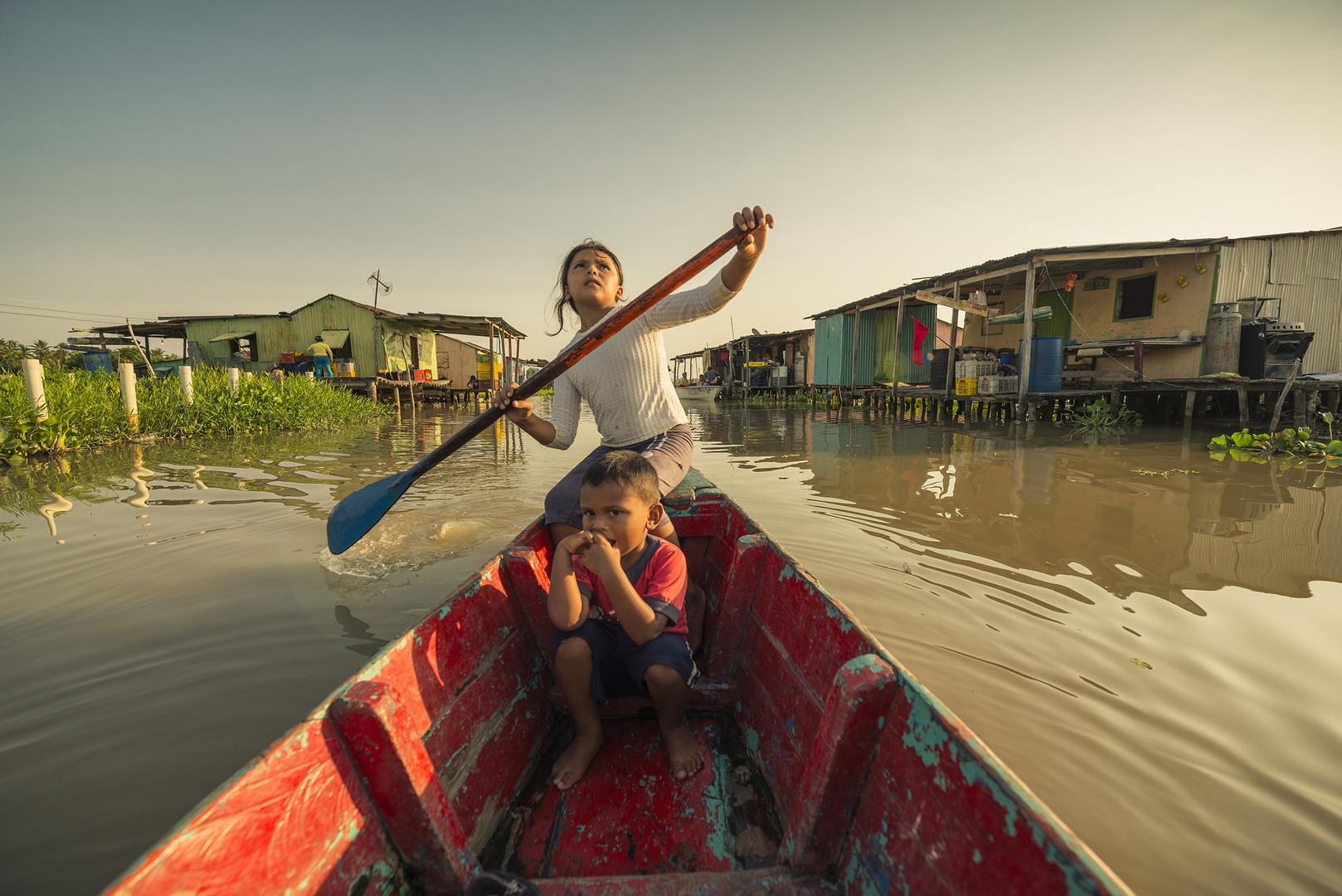 Still from Once Upon a Time in Venezuela Project: Hatched 2020. A little girl sitting with her brother rows a paddleboat through a community of houses built on the water.