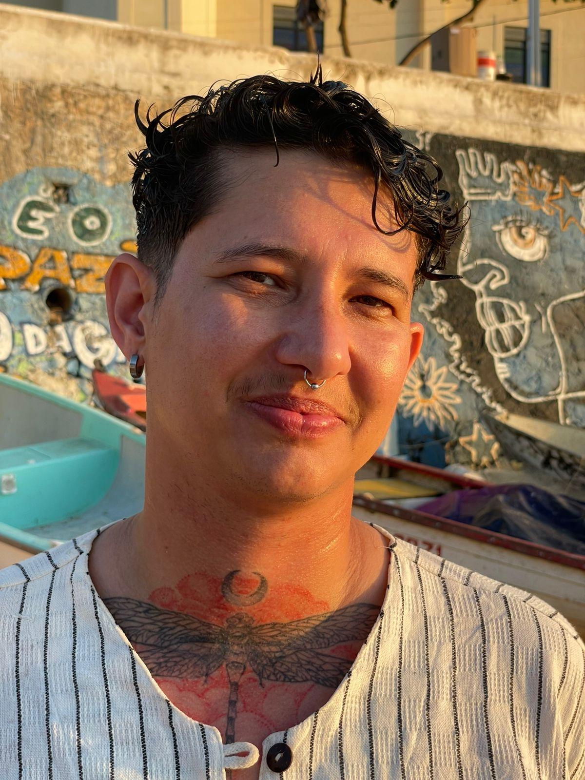 Headshot of a masculine presenting person smiling, with short black hair wearing a stripped grey and white shirt and a tattoo on his neck of a dragon-fly.