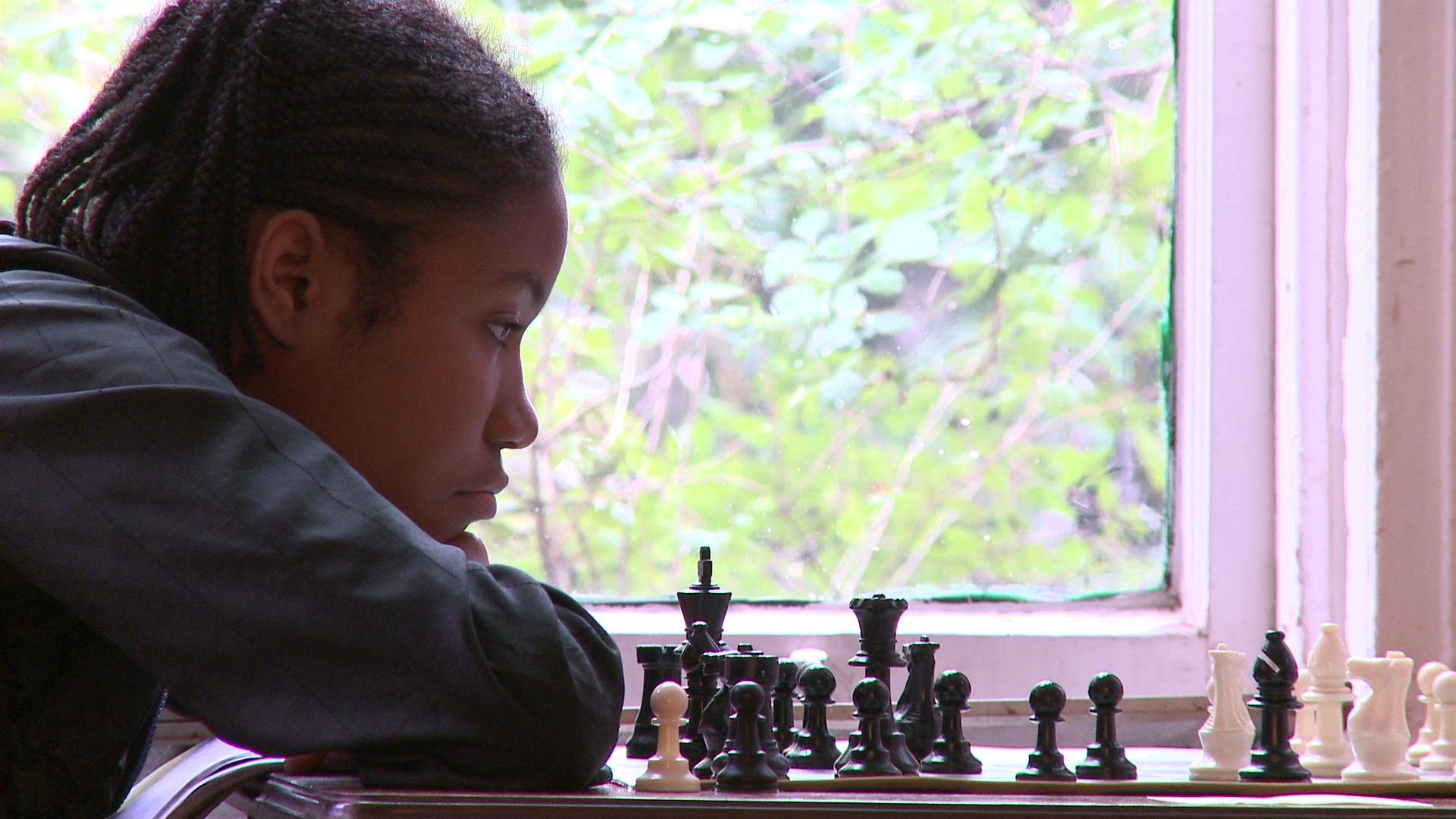 A young girl rests her head on her folded arms atop a chess board. She looks at the chess pieces.