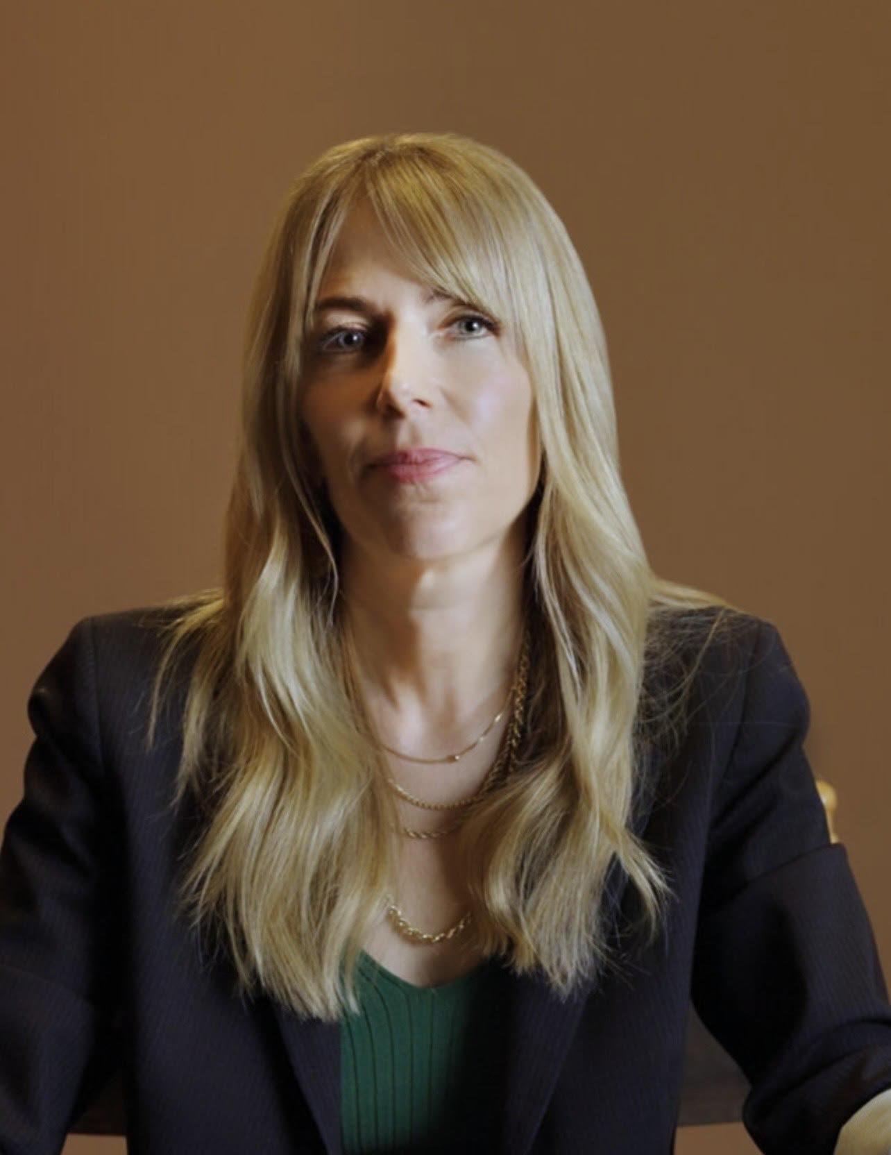 Headshot of Nancy Schwartzman wearing a black blazer in front of a brown background.
