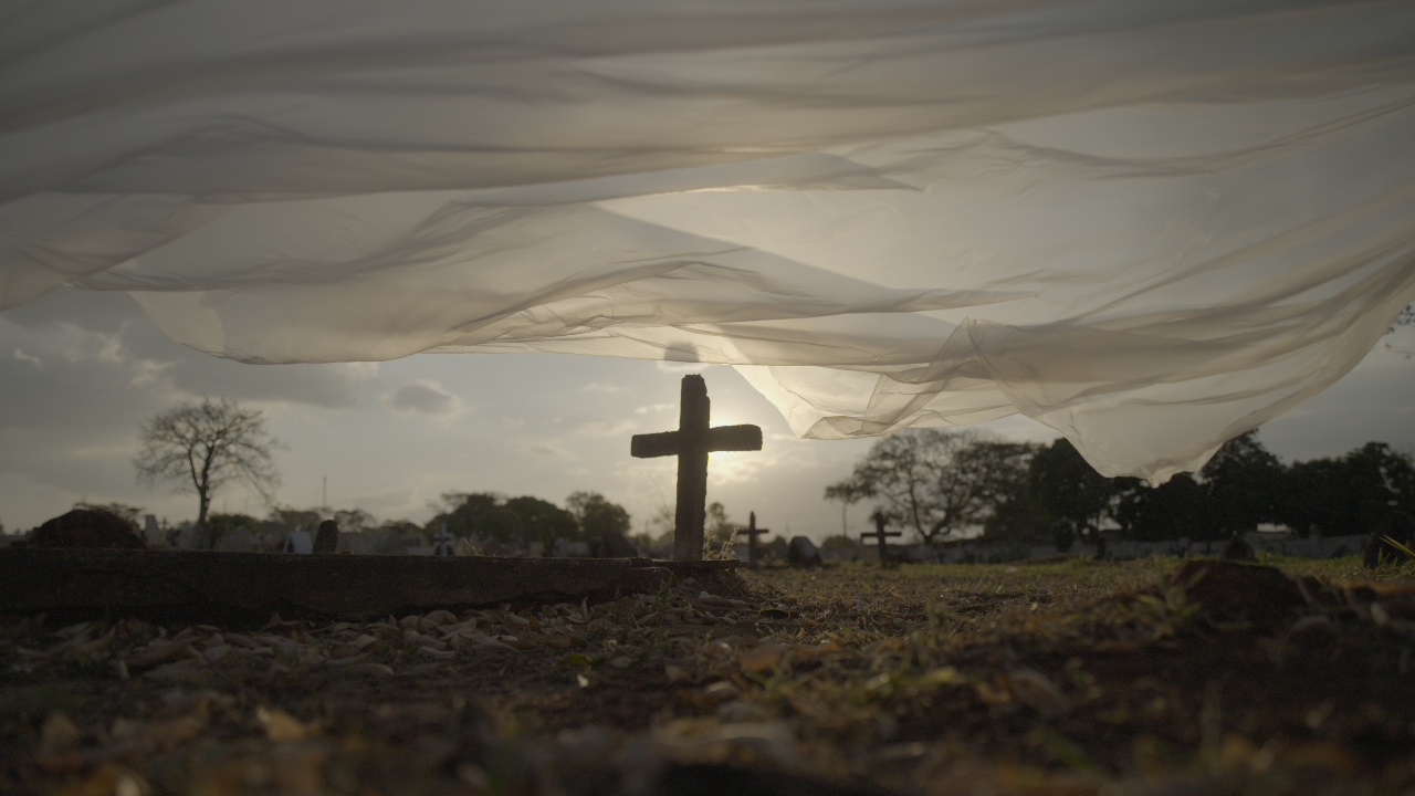 A statue of a cross is in the middle of a graveyard. There is a white sheet floating in front of it.