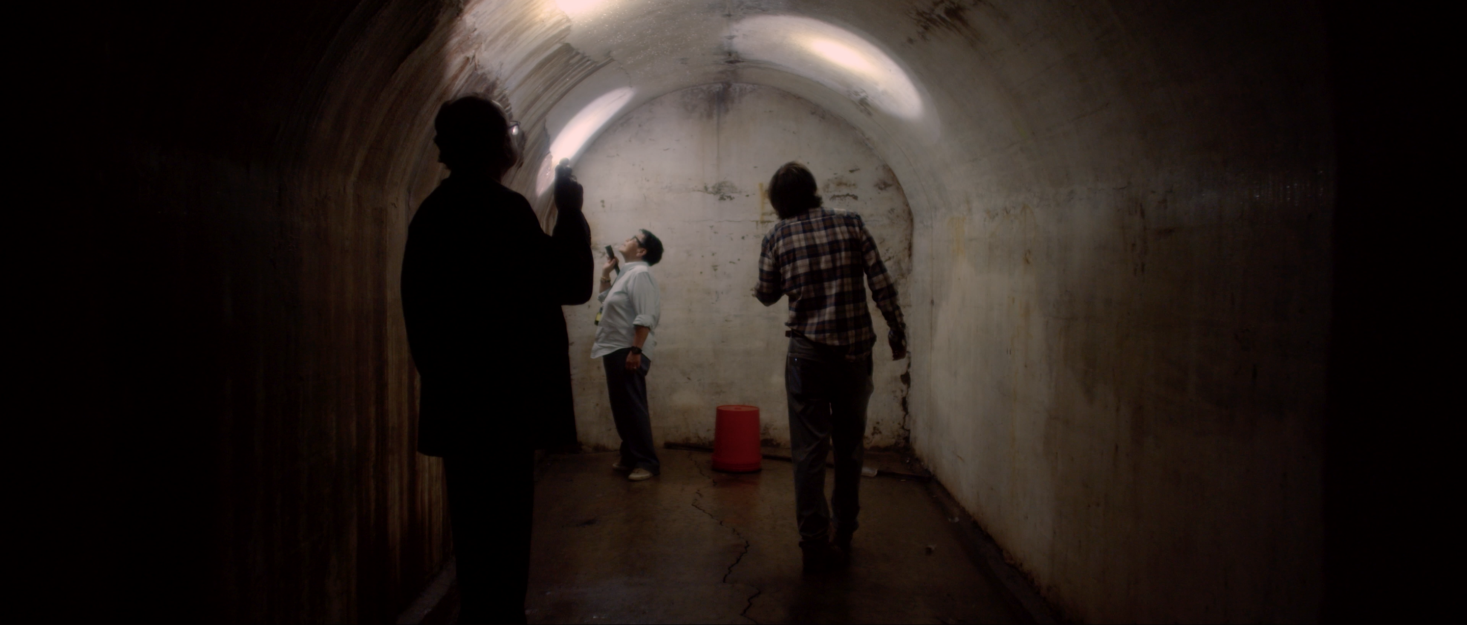 Inside a dark tunnel, three people point flashlights at the ceiling.