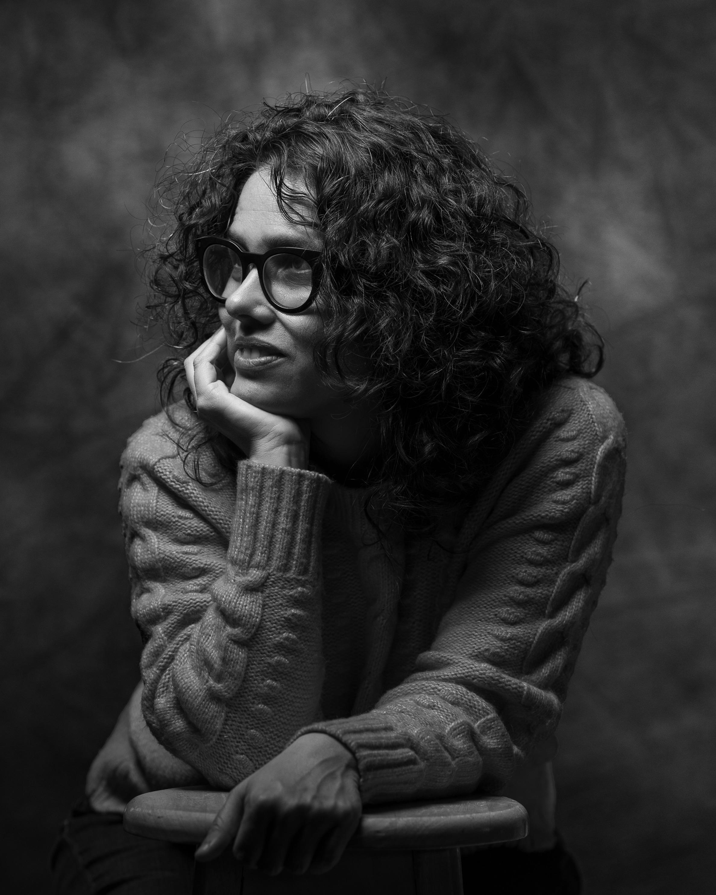 A black and white portrait of a woman resting her face on her hand and looking off camera. She wears glasses and has curly hair.