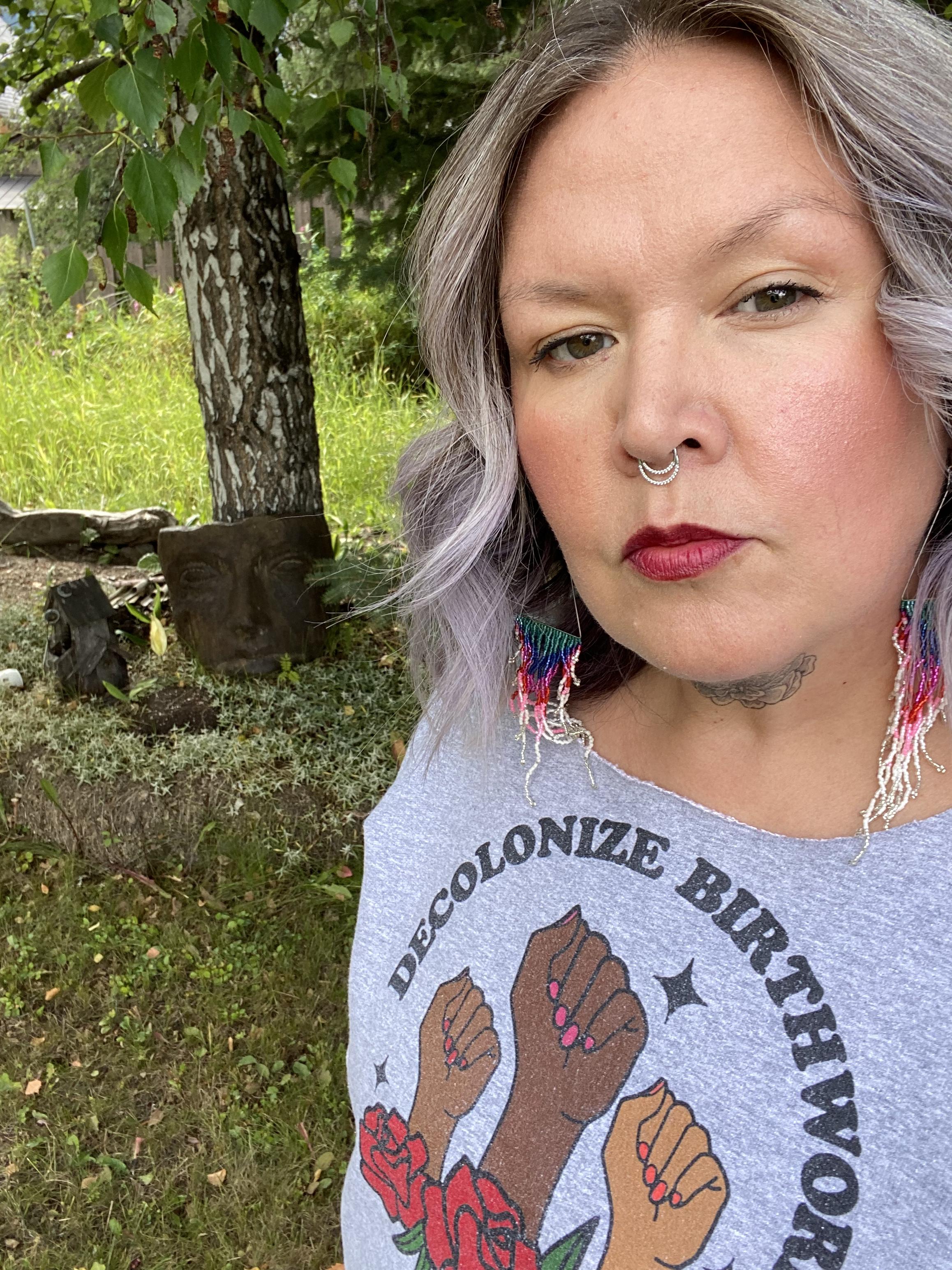 A person wearing beaded earrings in a grey shirt that reads "Decolonize Birthwork" looks at the camera with green grass and a tree behind them.