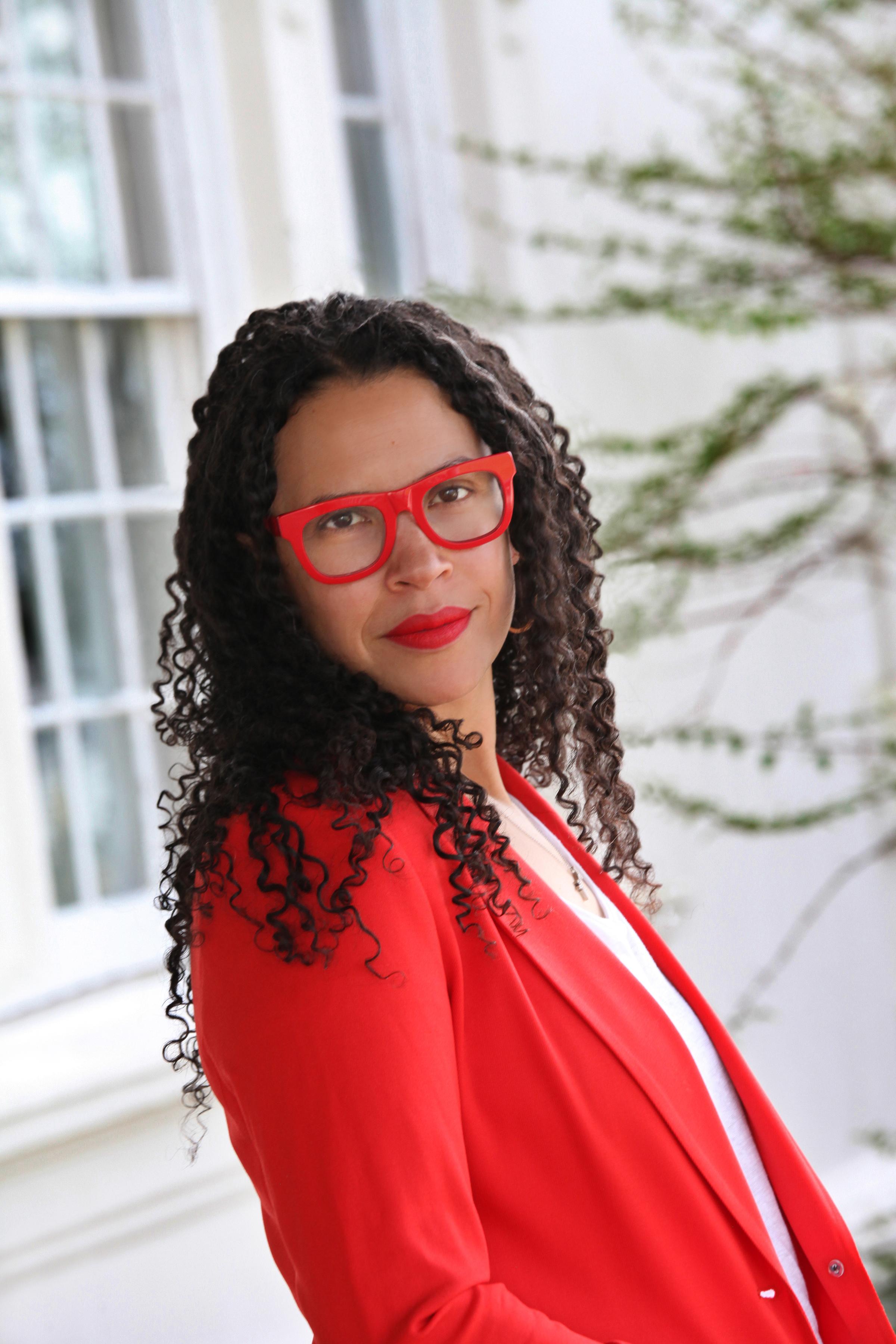 Light skinned black woman with long curly hair wearing red glasses and a red jacket.